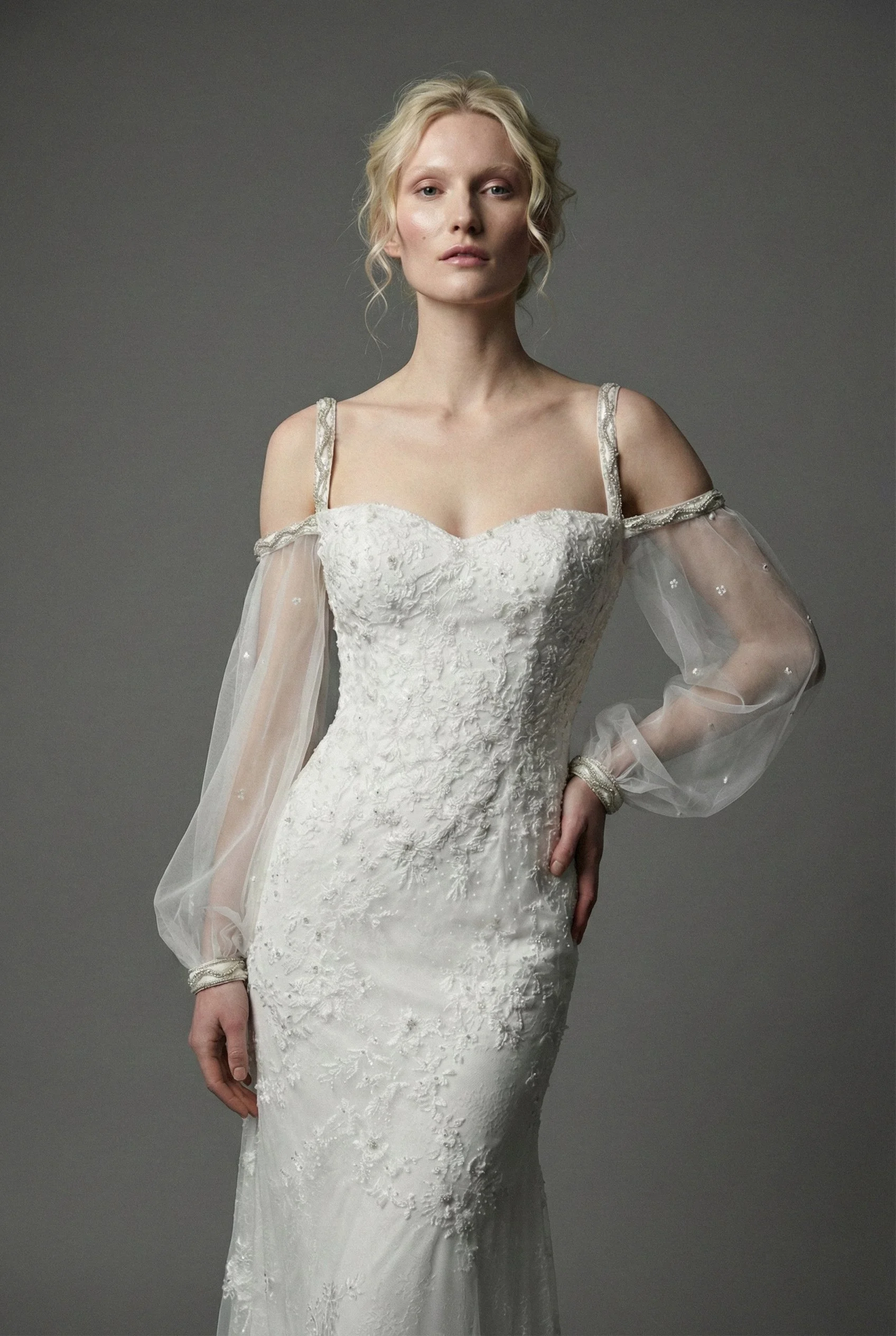 A woman in a white Tess Mann Atelier luxury wedding dress with lace and embroidery, sheer long sleeves, and shoulder straps, standing against a gray background.