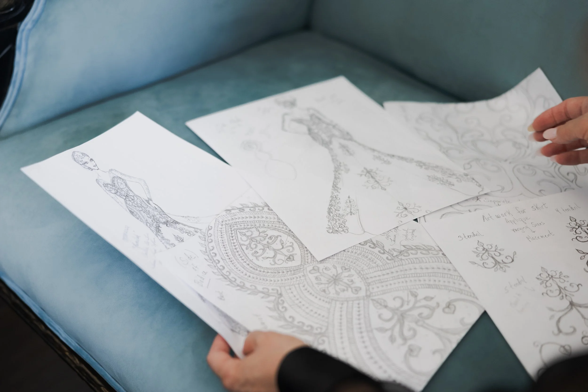 Sketches of fashion designs featuring dresses and intricate patterns on white paper, with a person's hand holding one of the sketches.