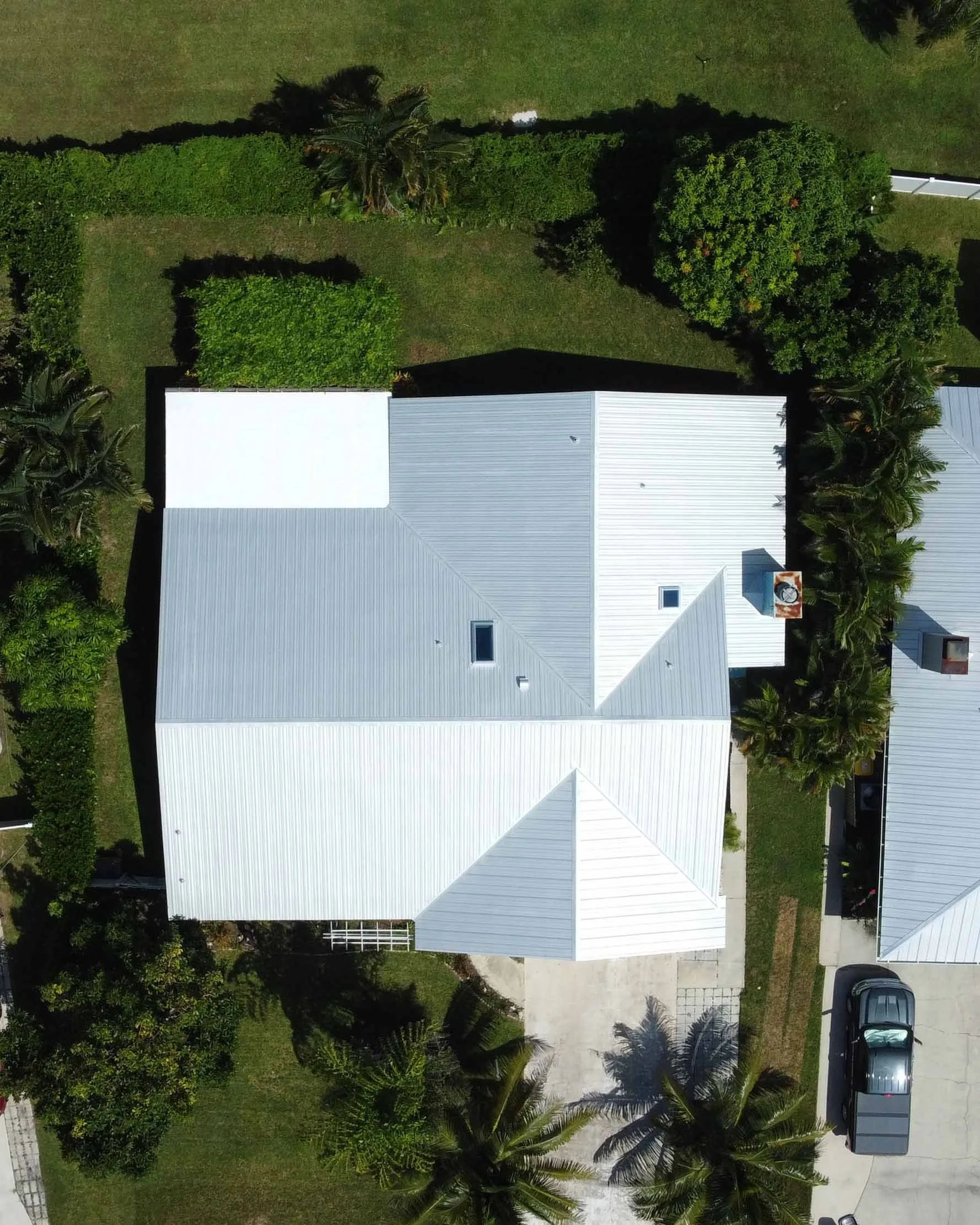 Aerial view of a house with a white metal roof, surrounded by greenery and trees, with a driveway and parked car visible.