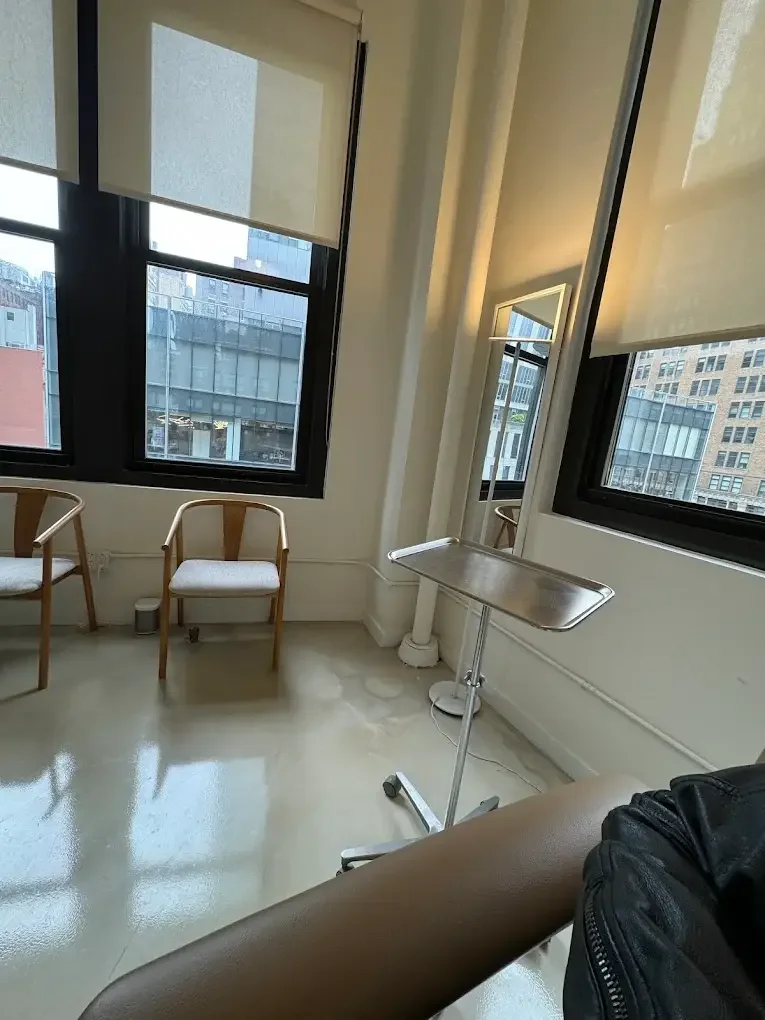 A woman lying on a bed receiving a cosmetic treatment on her nose from a technician in a bright room with large windows and city buildings outside.