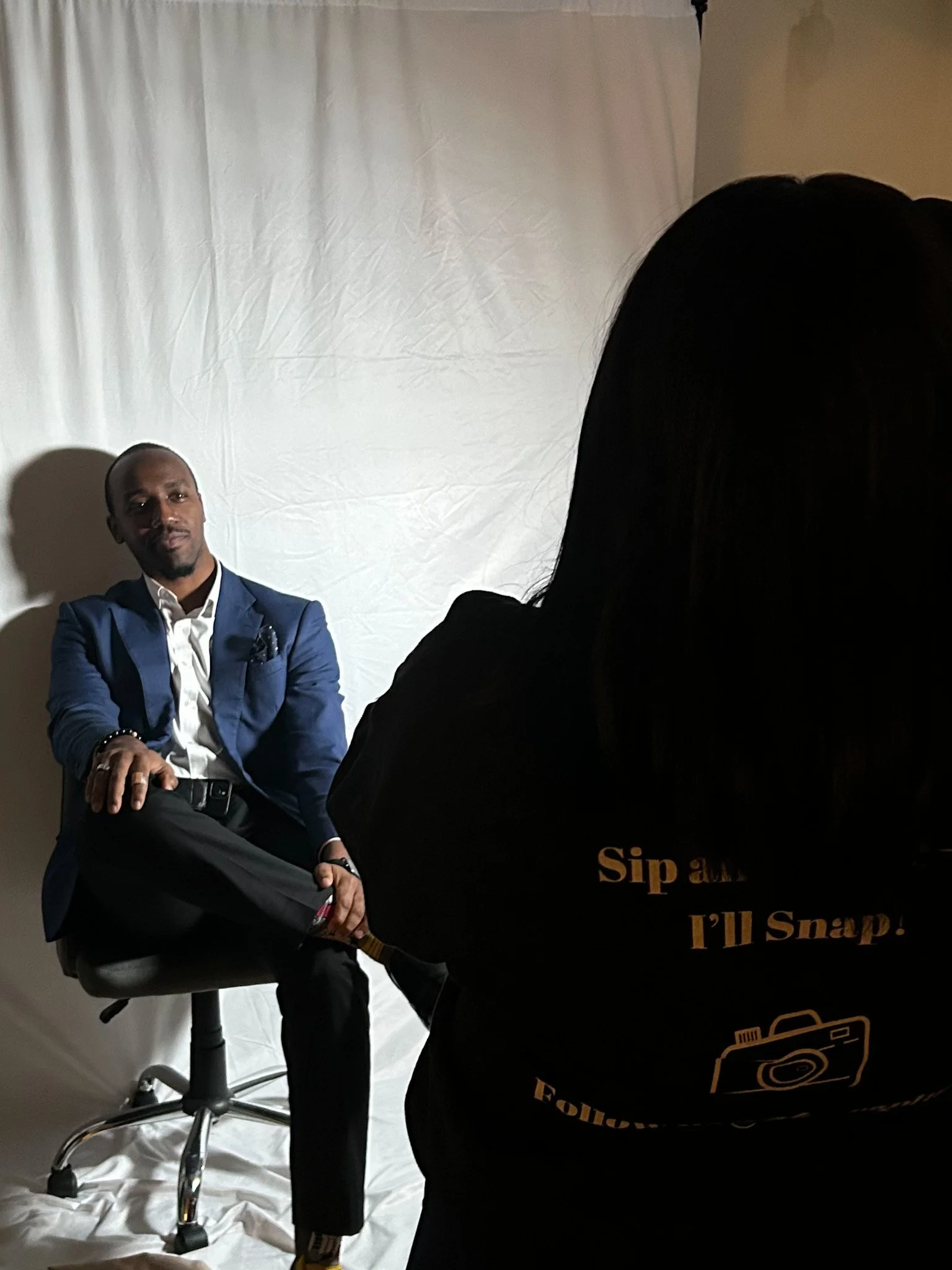 Man in a blue suit and white shirt sitting on a stool, looking at the camera, with a person taking a photo in the foreground. The background is a white curtain.