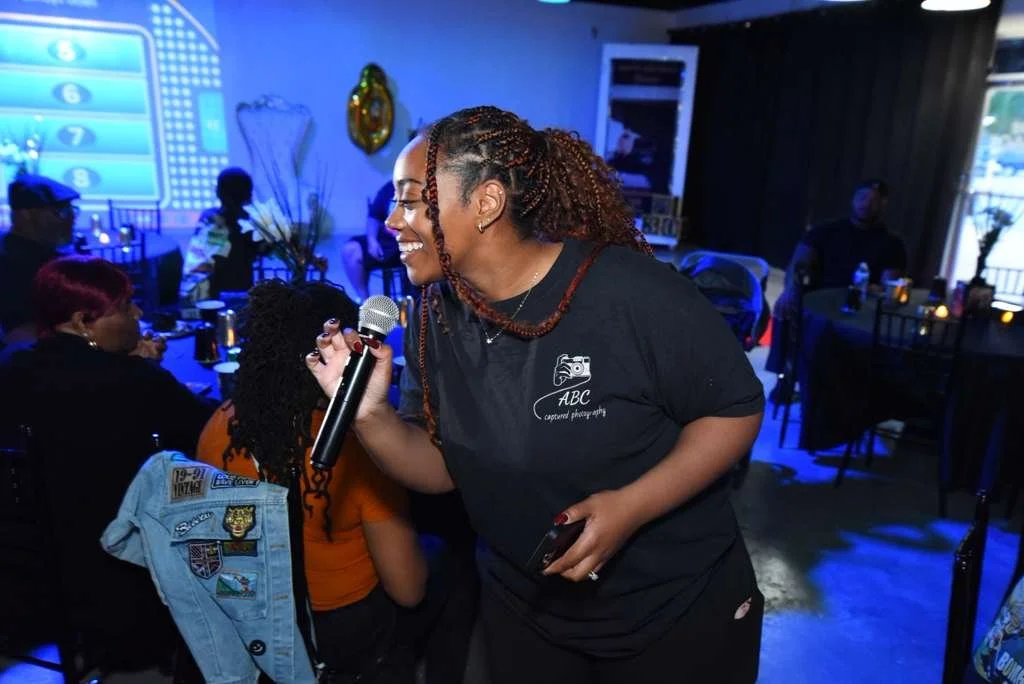 A smiling woman with braided hair holding a microphone and talking to a person in an orange shirt at a gathering in a dimly lit room with blue lighting, decorated with balloons and tables.
