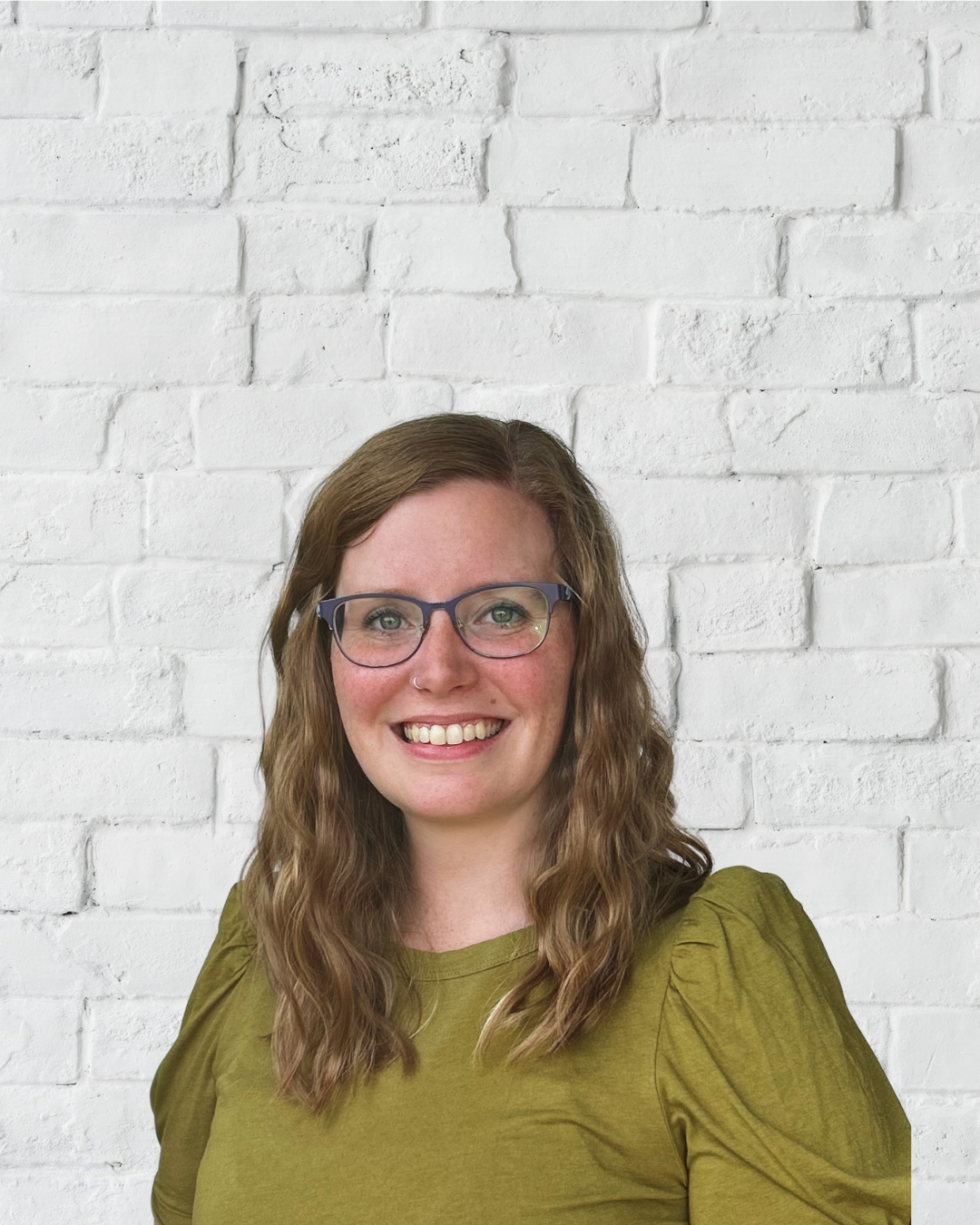 A woman with wavy brown hair, glasses, and a nose ring smiling in front of a white brick wall.