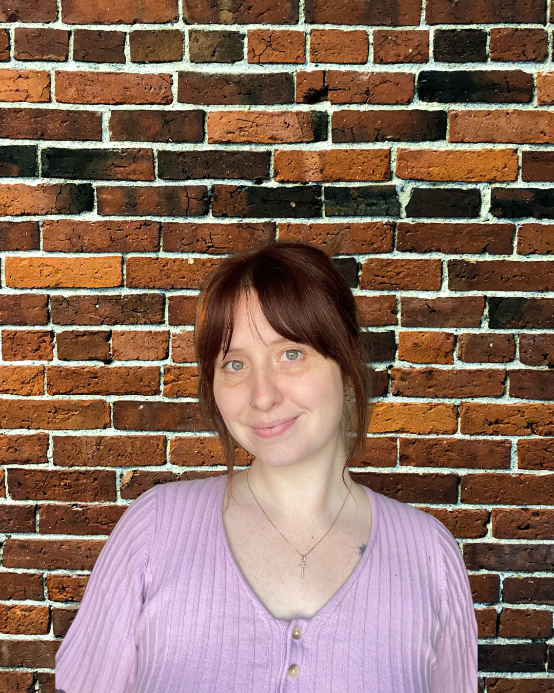 A woman with reddish-brown hair and light skin smiling in front of a red brick wall. She is wearing a light purple top with buttons and a silver cross necklace.