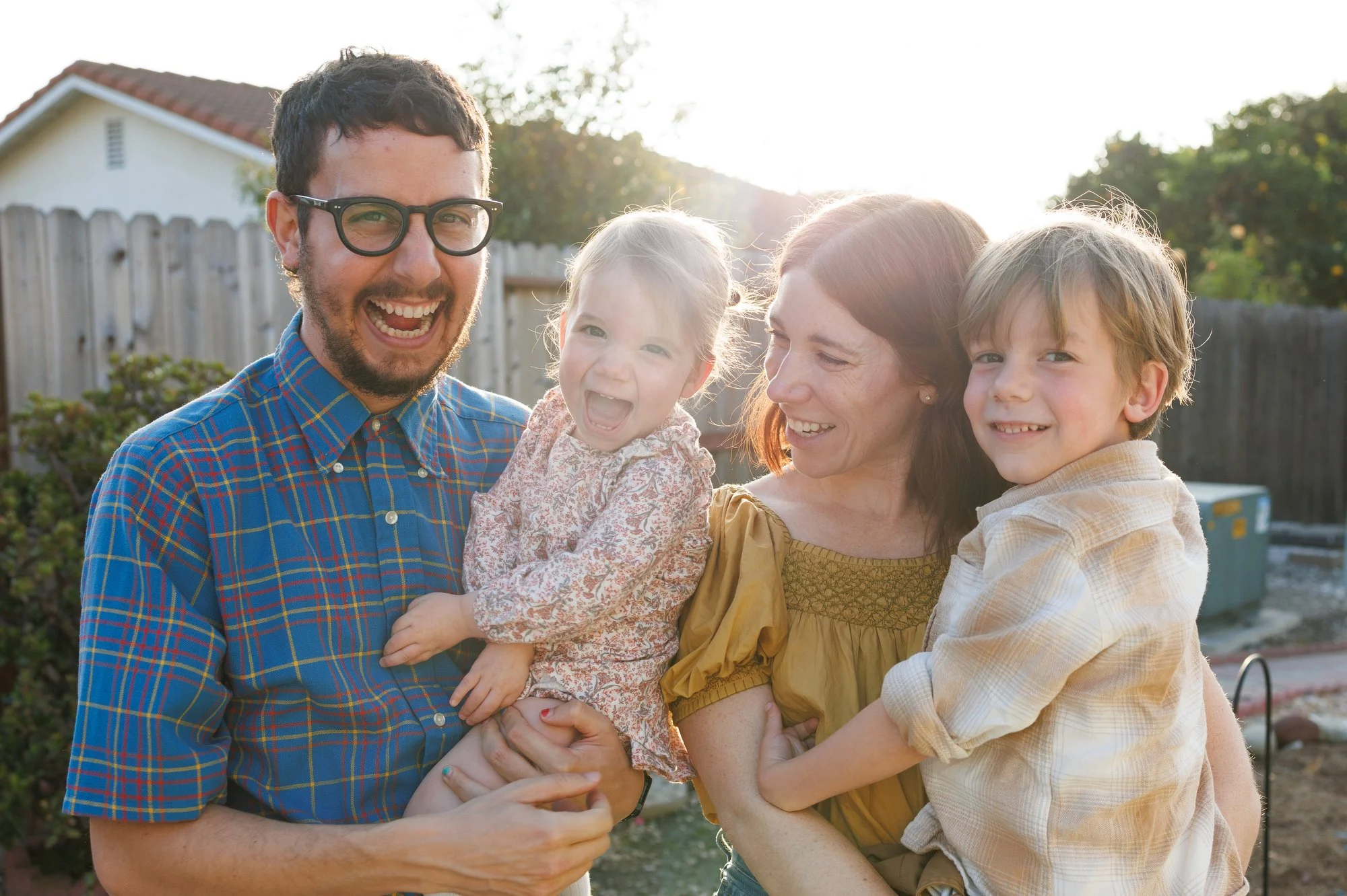 A dad holds his daughter and a mom holds her son, all laughing, with bright sun behind them