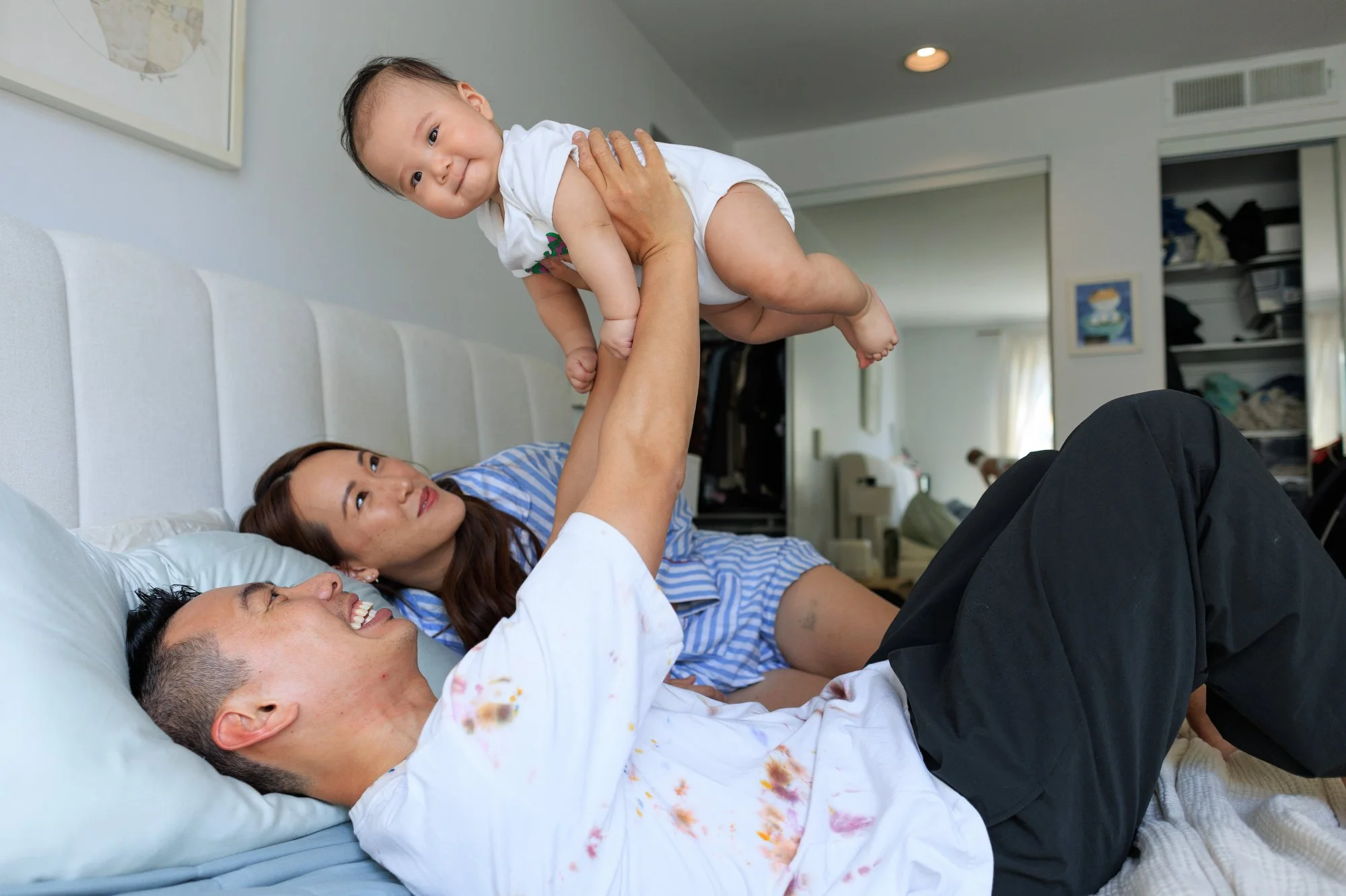 holiday card photographer: a dad holds his baby above his head while laying on the bed. mom looks on and smiles. baby smiles at the camera.