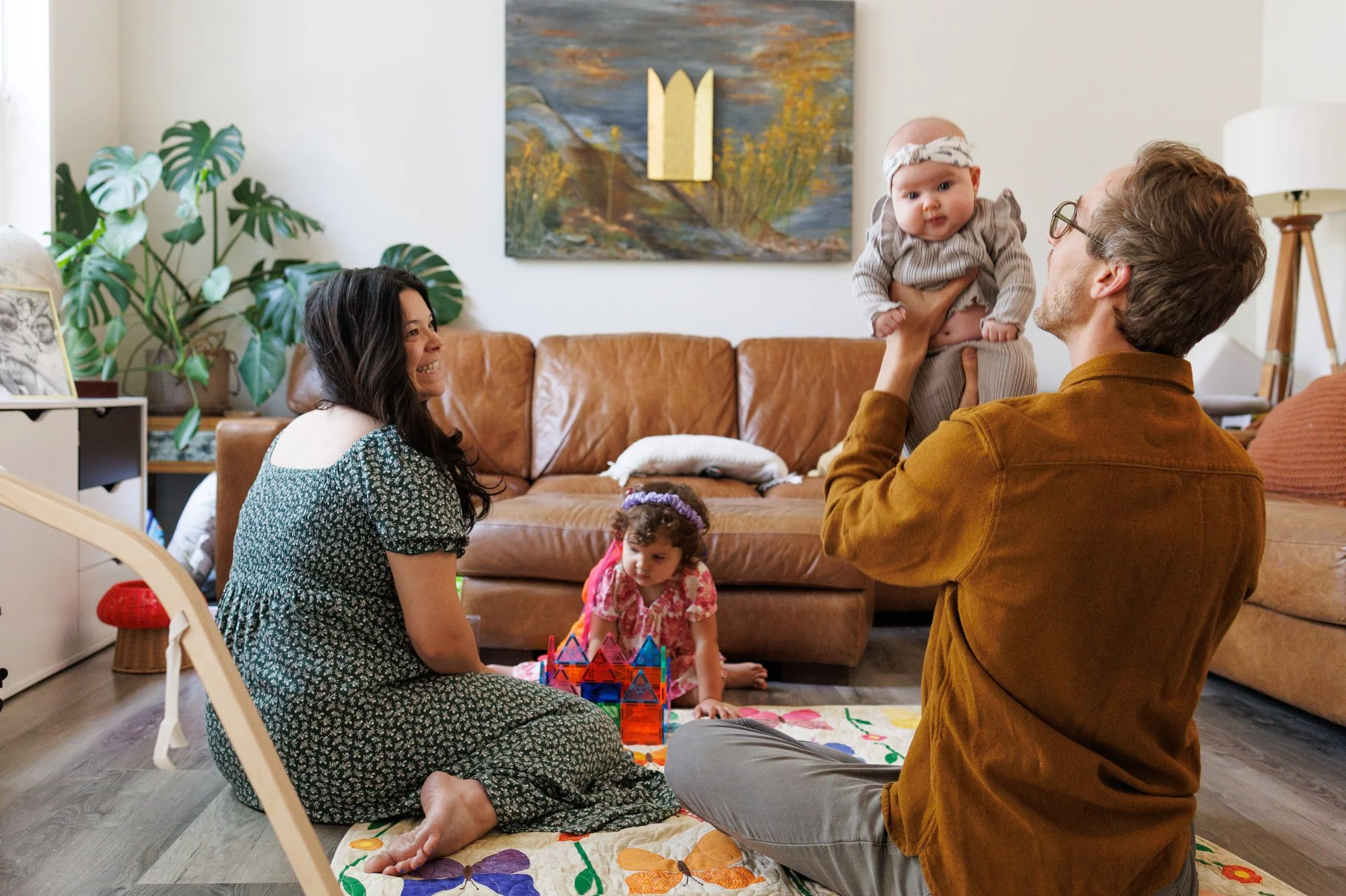 Average cost of family photos: Dad holds his baby up while mom looks on smiling. A little girl plays with Magna-Tiles on the floor. A landscape painting hangs in the background.