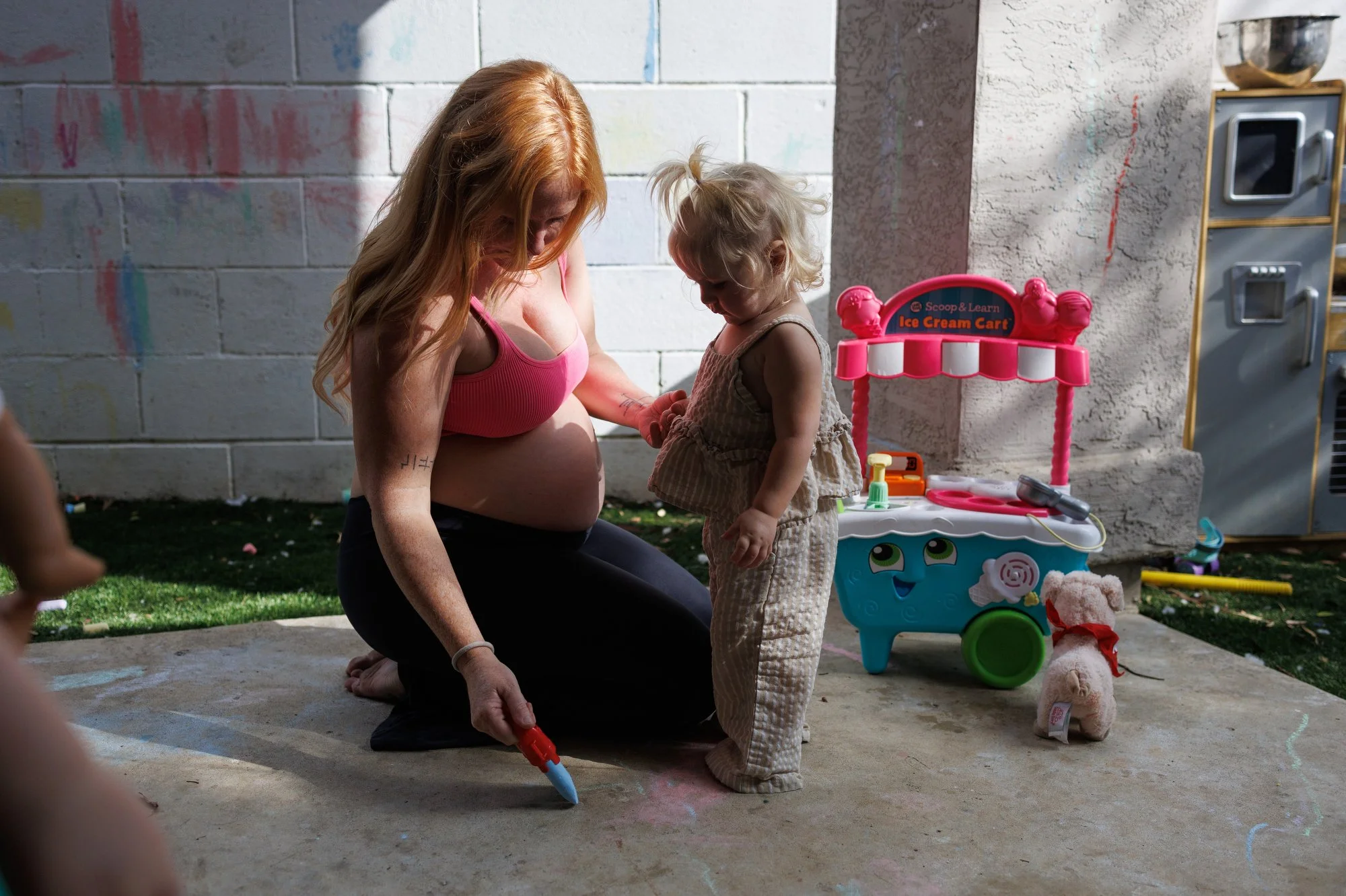 A pregnant mom holds toddler's hand as she draws with chalk on concrete. Backyard toys are scattered around them. Photo by OC family photographer Shannon Fisher.