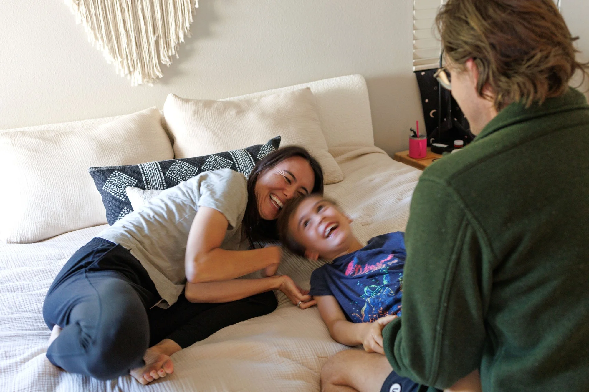 Family photoshoot outfit ideas: A mom and son laugh on the bed as the dad holds his son's hands. Mom wears a grey T-shirt and black sweats. Son is in graphic tee. Dad is in a dark green structured shirt.