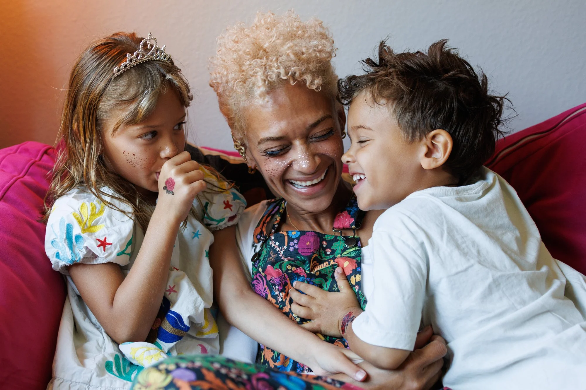 Family photoshoot outfit ideas: Mom wears floral print overalls. Little girl wears a tiara and white dress with colorful appliqué. Son in white shirt. All have temporary tattoos and face glitter.