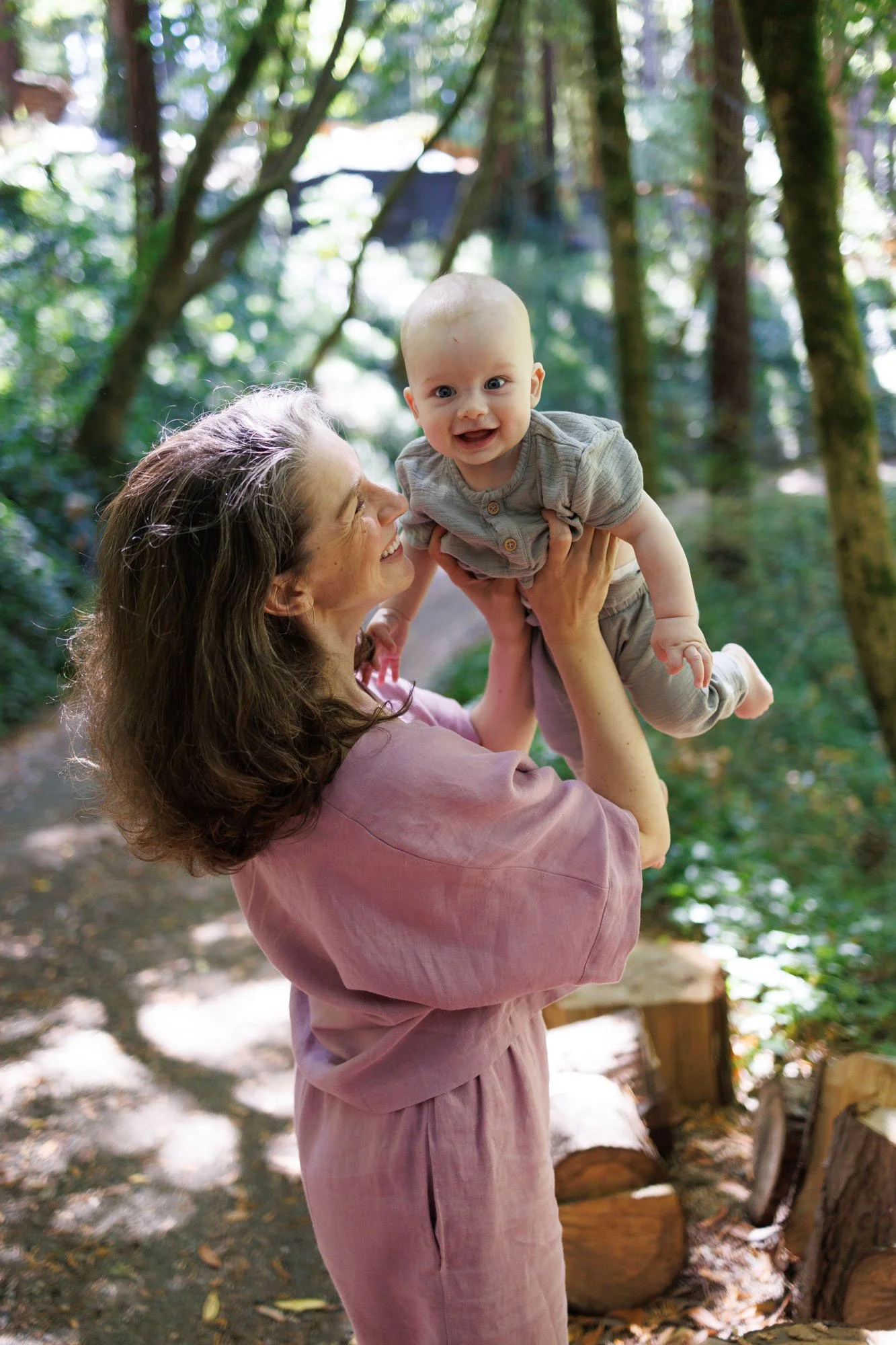 Mom in a dusty mauve top and pants holds her baby up in the air as he smiles and looks in the camera. They are on a forest path.