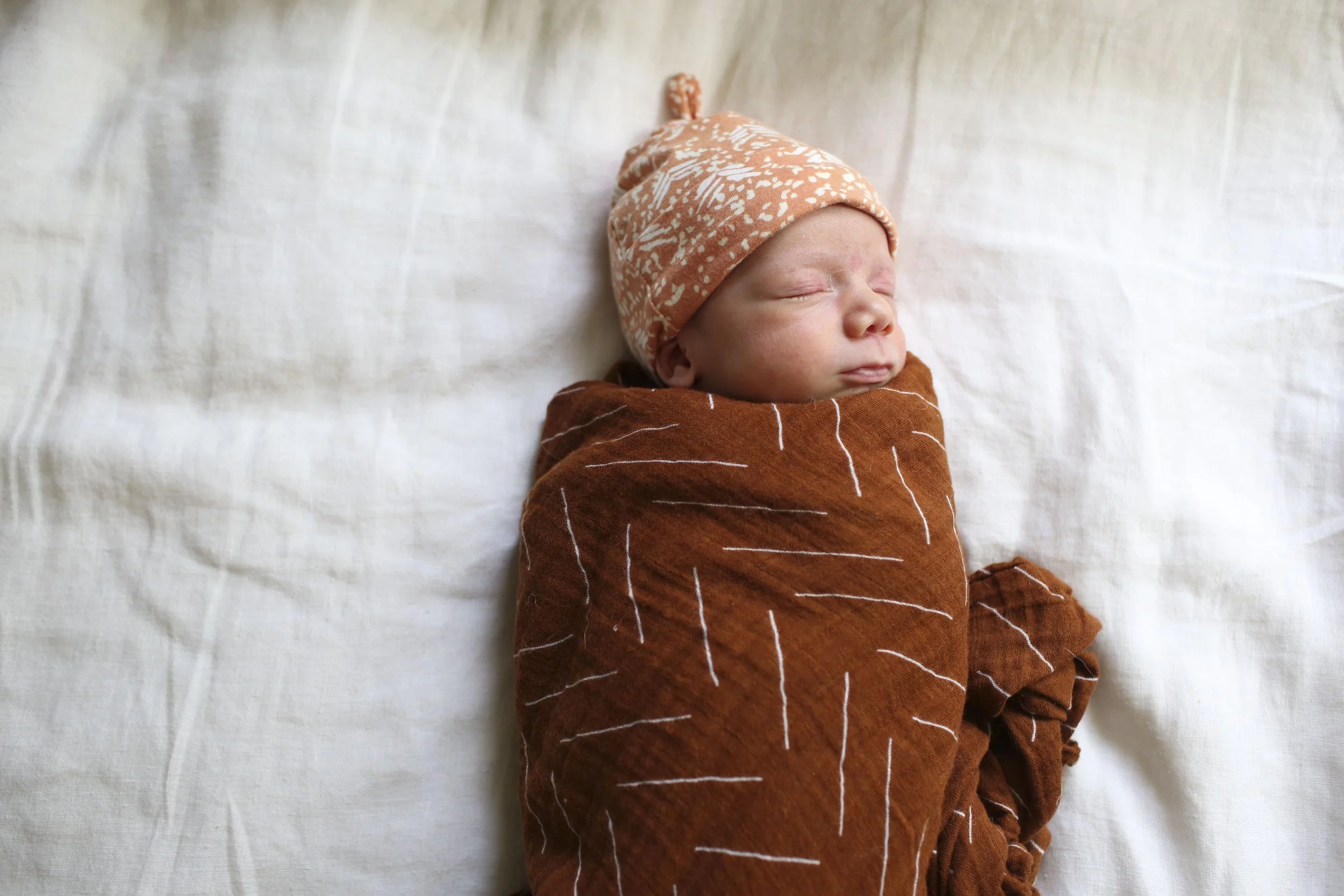 Baby closes his eyes while swaddled in a rust-colored muslin cloth