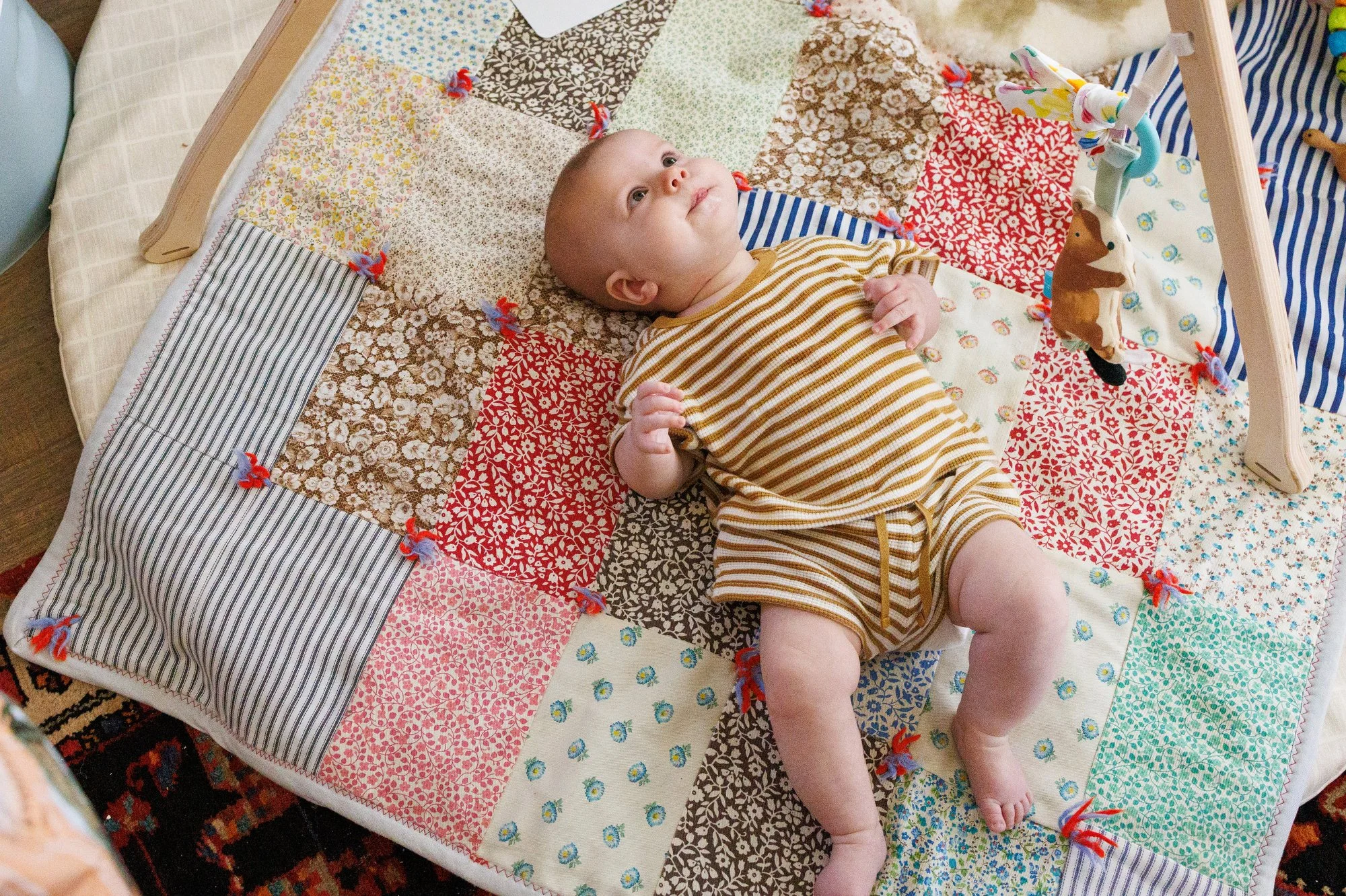 A newborn in a striped shirt and shorts lays on his back on a patchwork quilt. He is looking up at something offscreen. Photo by Shannon Fisher, newborn photographer in Orange County.