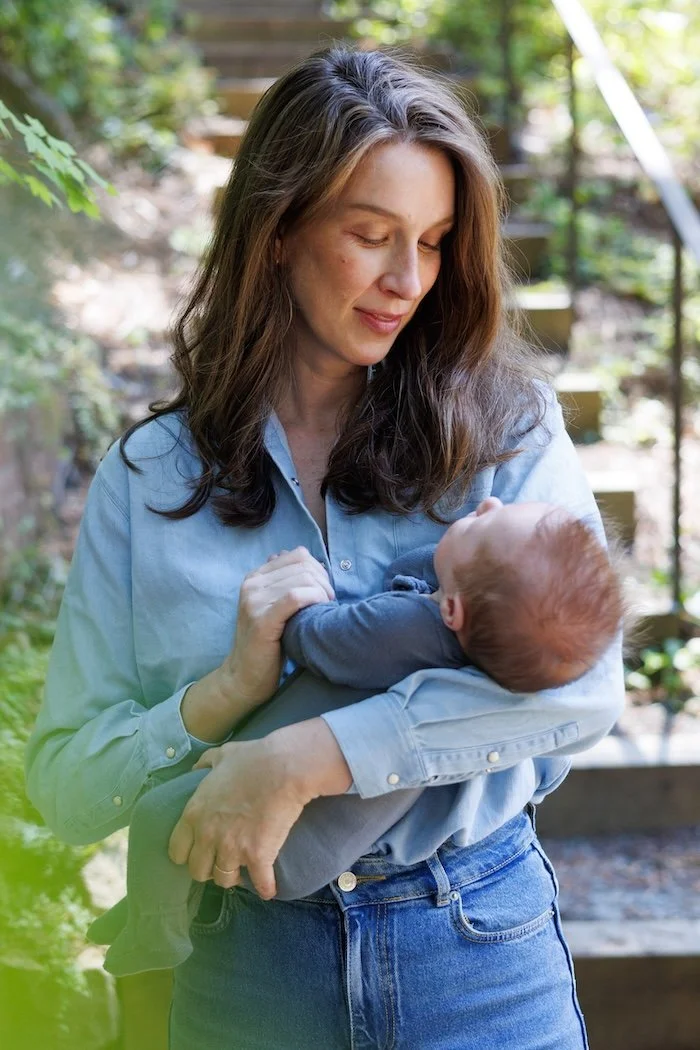Average cost of family photos: A mom in a blue button-up looks in her baby's eyes as she holds him. There's a green lens flare in the bottom left corner.