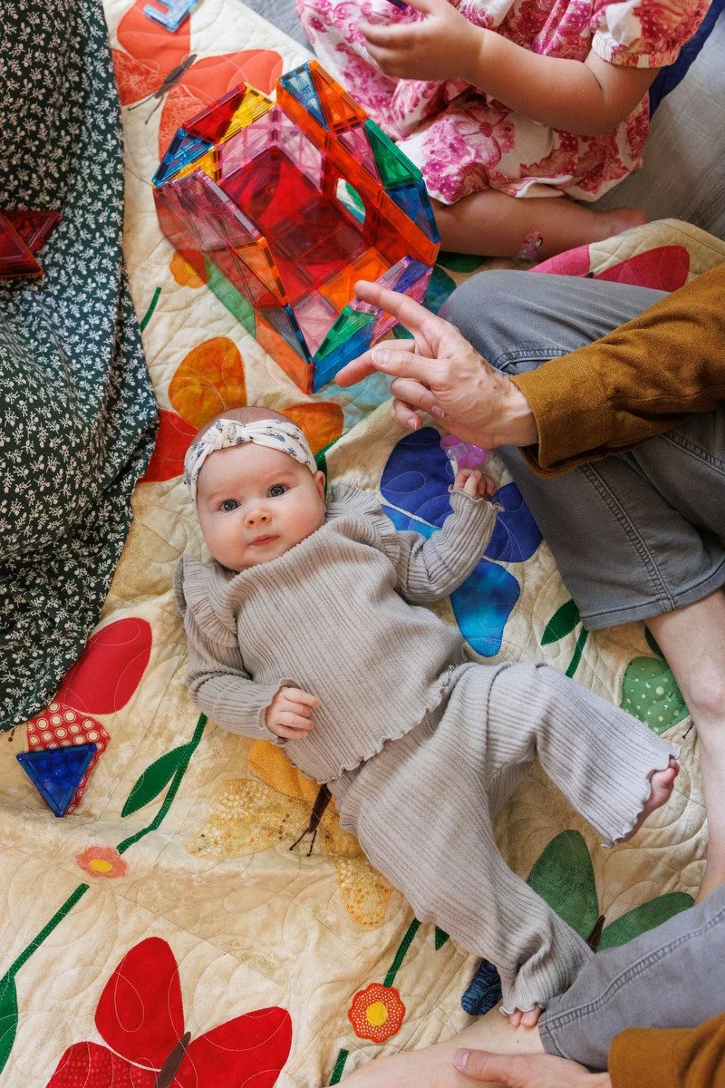 Baby in grey ruffled pants and shirt with a headband lays on blanket with adults' legs peeking into the shot. Photographer stands straight over her as she looks into the lens.