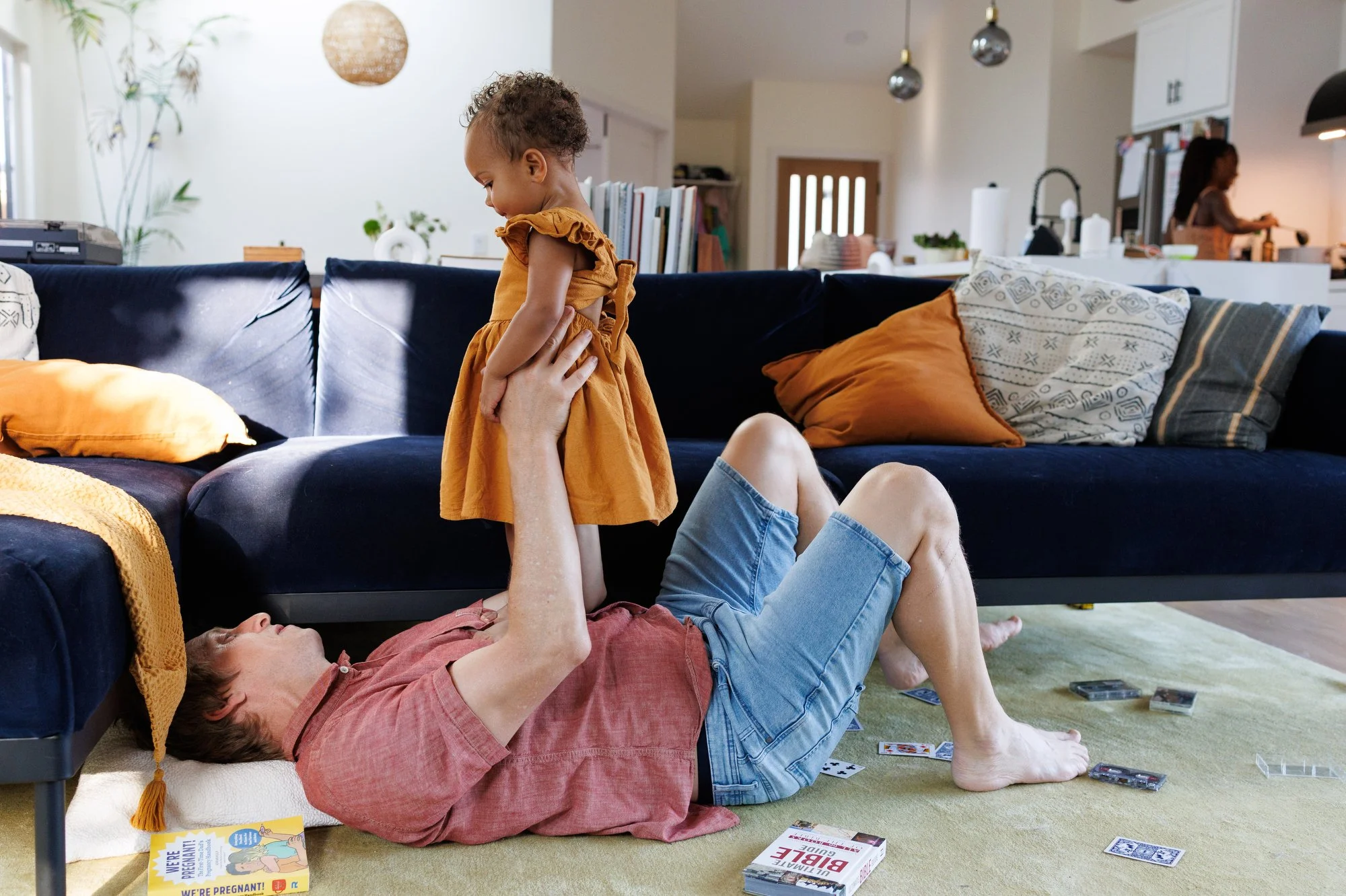 In a documentary-style photo session at home by Shannon Fisher, a toddler in a yellow dress stands on her dad's stomach in their living room.
