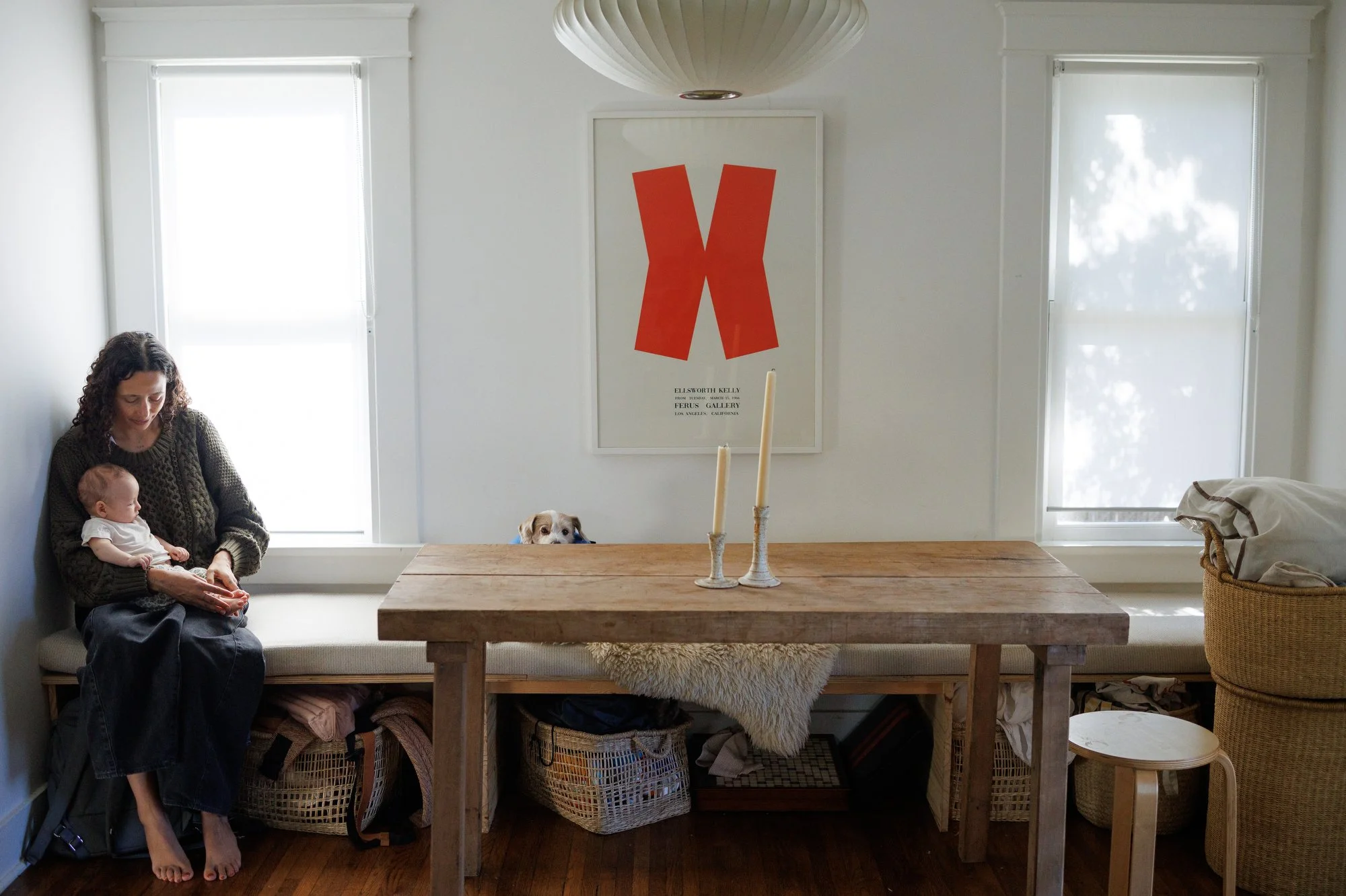 Mom holds baby in her lap sitting on a bench next to a rustic wood dining table. A minimal print with a red X shape hangs on the wall. A dog peeks above the table. OC newborn photography by Shannon Fisher.
