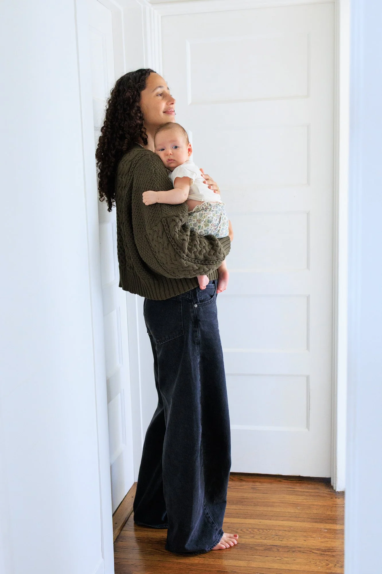 Family photoshoot outfit ideas: Mom holds baby to her chest, smiling softly. She wears an olive sweater and wide-leg black jeans. Baby wears olive and blue floral pants and white shirt.