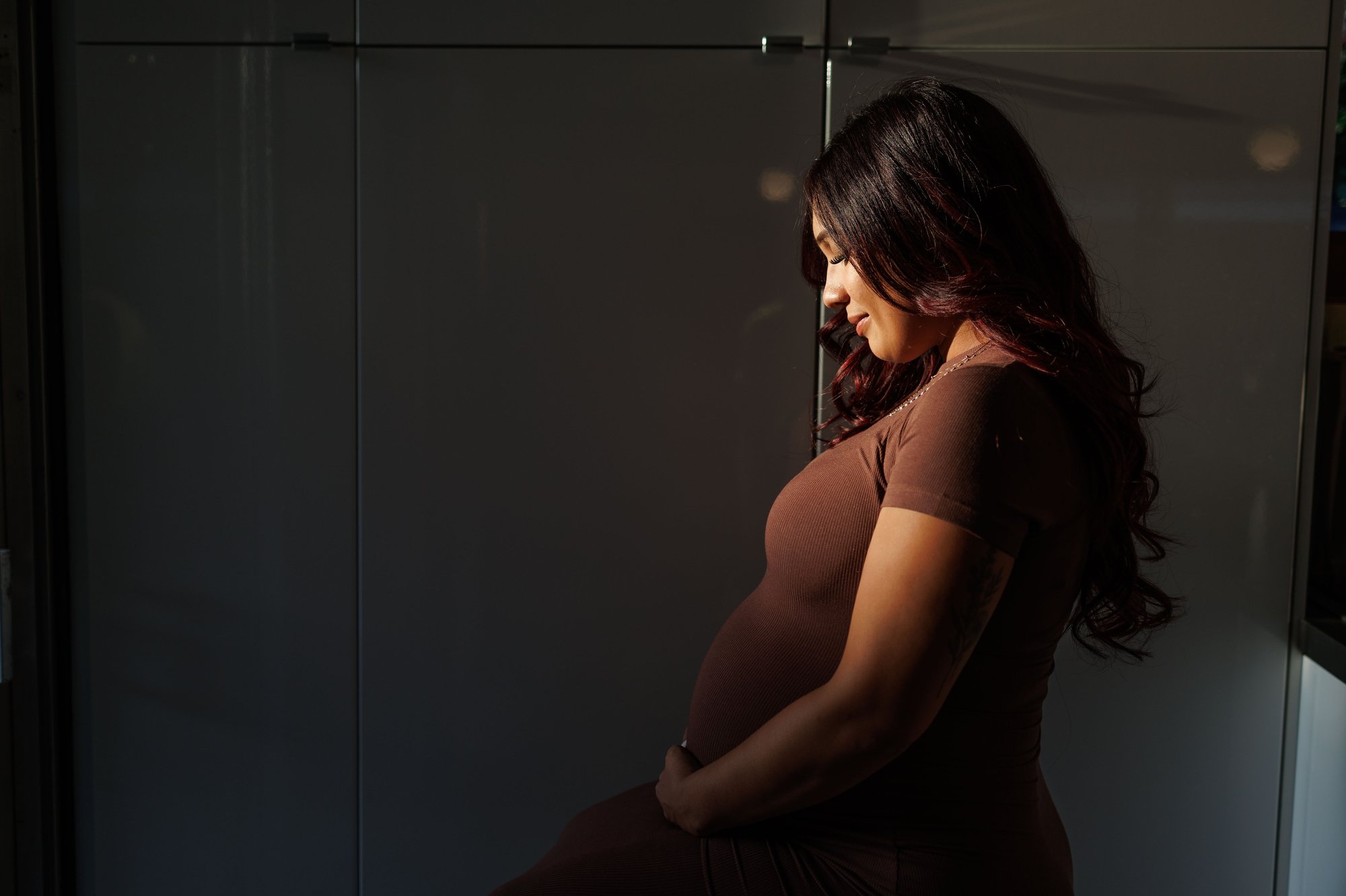 orange county maternity photographer: a woman in a brown dress holds her pregnant belly as she looks down with sun illuminating her face. photo by shannon fisher.