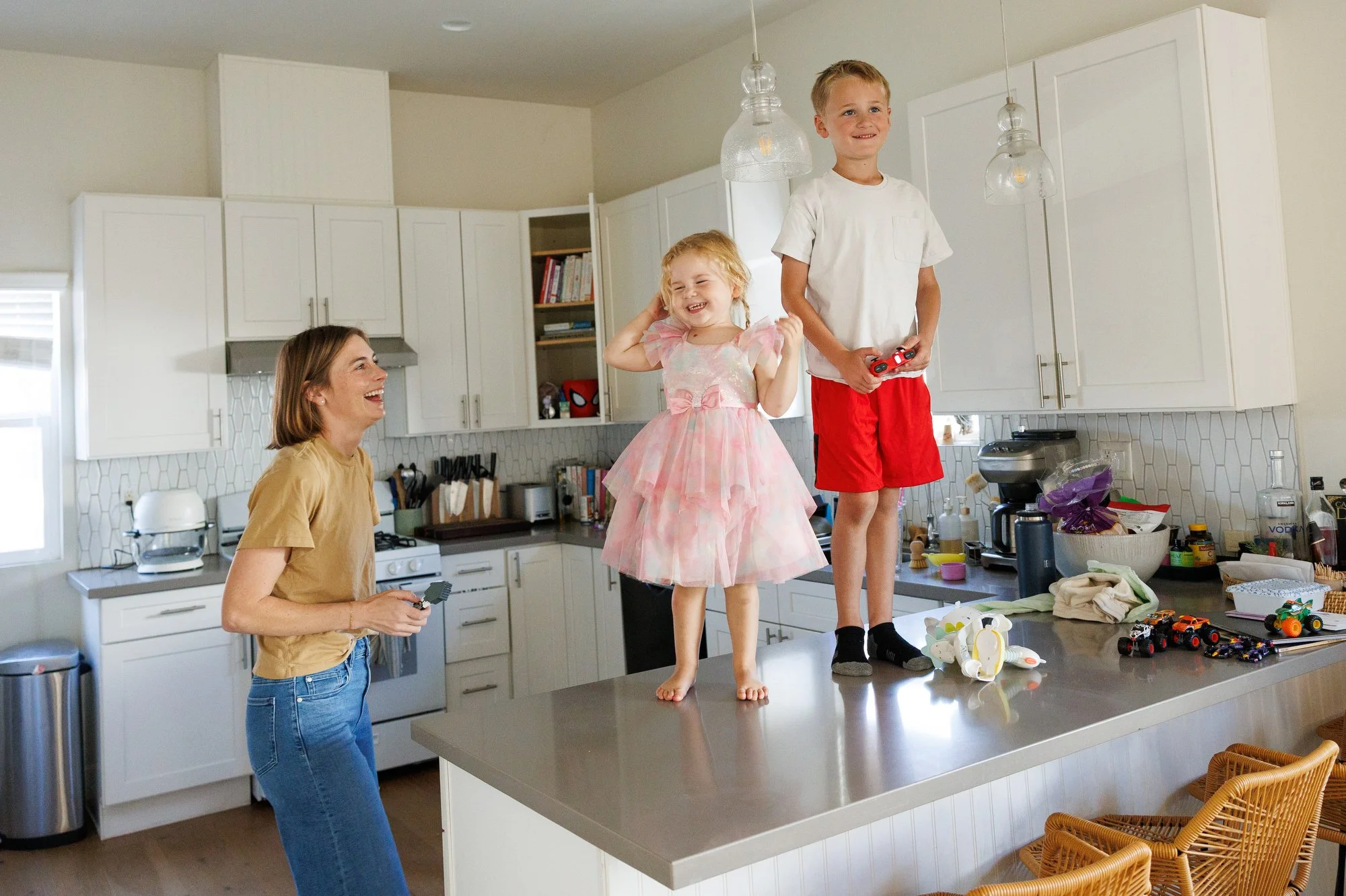 A little girl in a ruffled pink dress laughs while standing on a kitchen countertop. Her brother in red shorts stands beside her. Mom looks on, laughing. Photo by Orange County lifestyle family photographer Shannon Fisher.