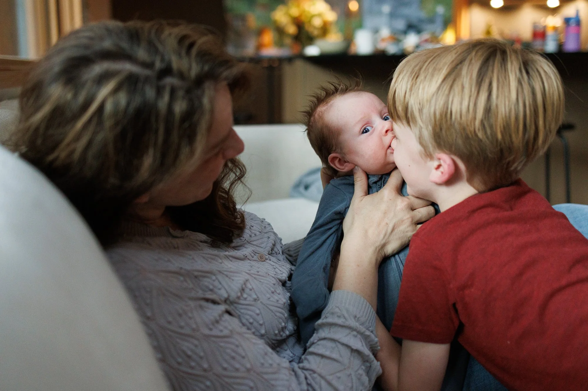 Baby with hair sticking straight up looks into the camera while biting brother's nose. Mom is holding baby up.