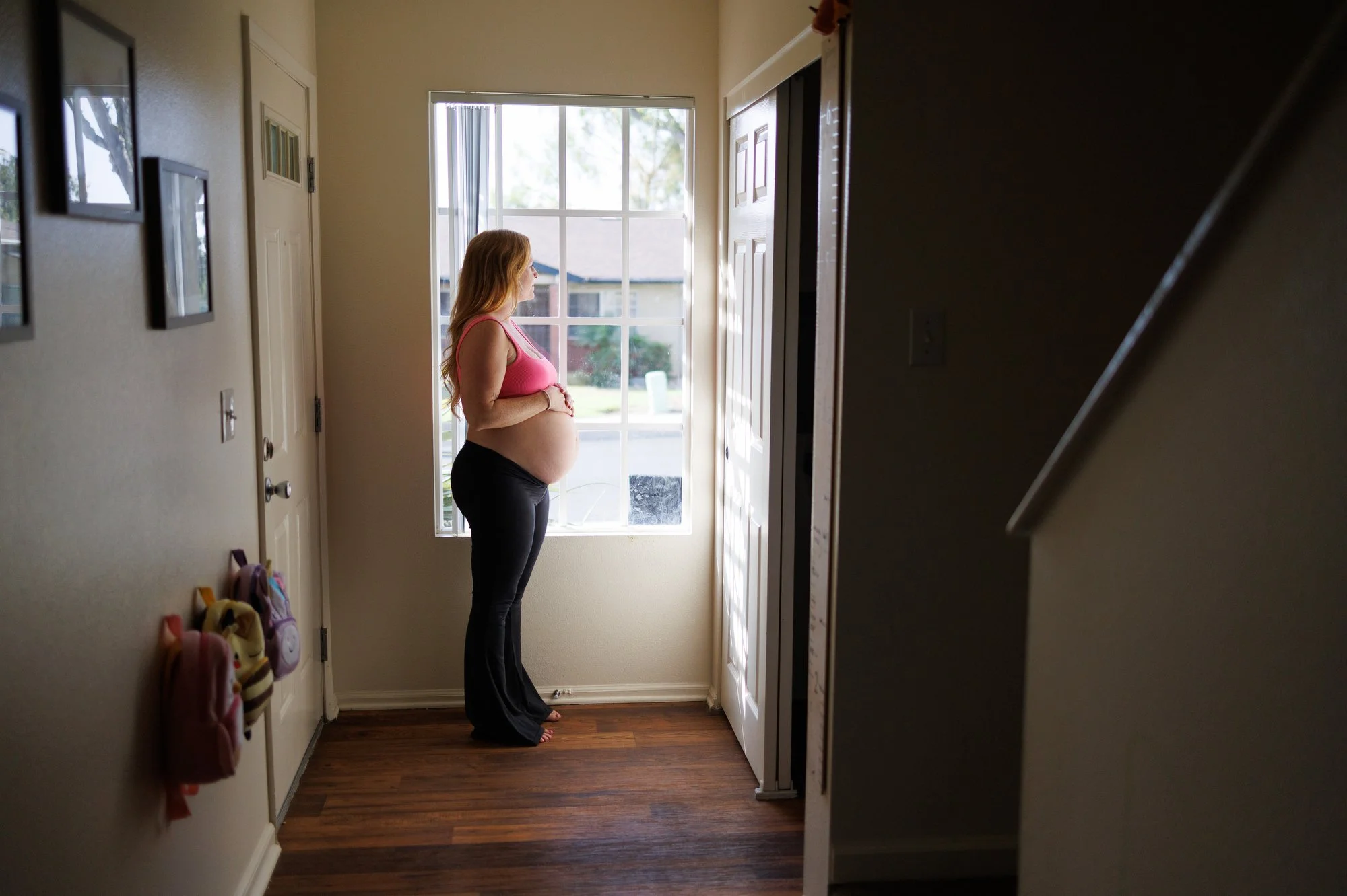 a woman in wide-legged yoga pants and a pink sports bra stands by the window holding her pregnant belly