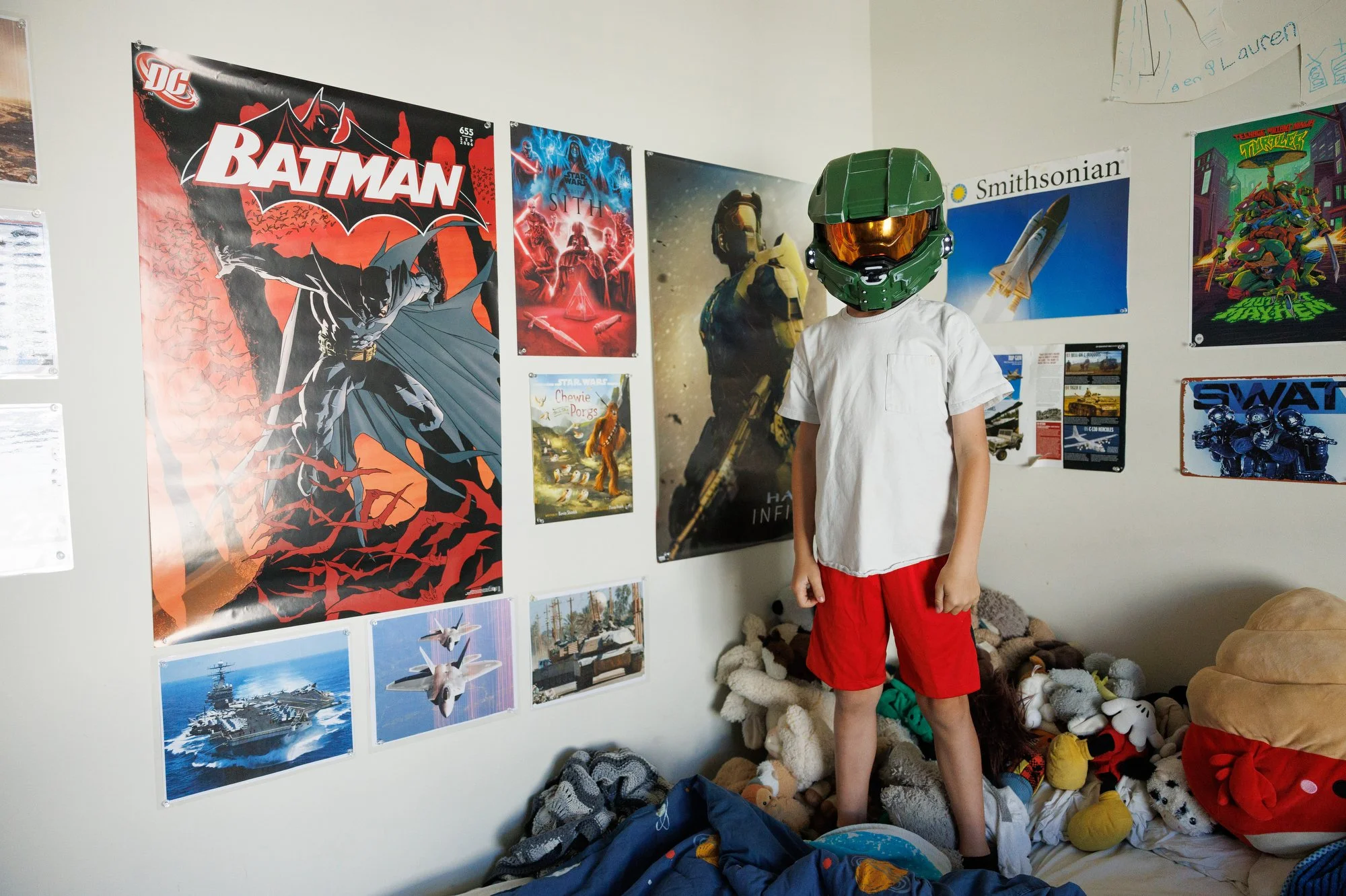 A boy in a green Halo helmet with a copper face shield stands on his bed, surrounded by stuffies. His walls are covered in posters of planes, comics, and rockets. Photo by Shannon Fisher, a documentary-style in-home family photographer in OC.