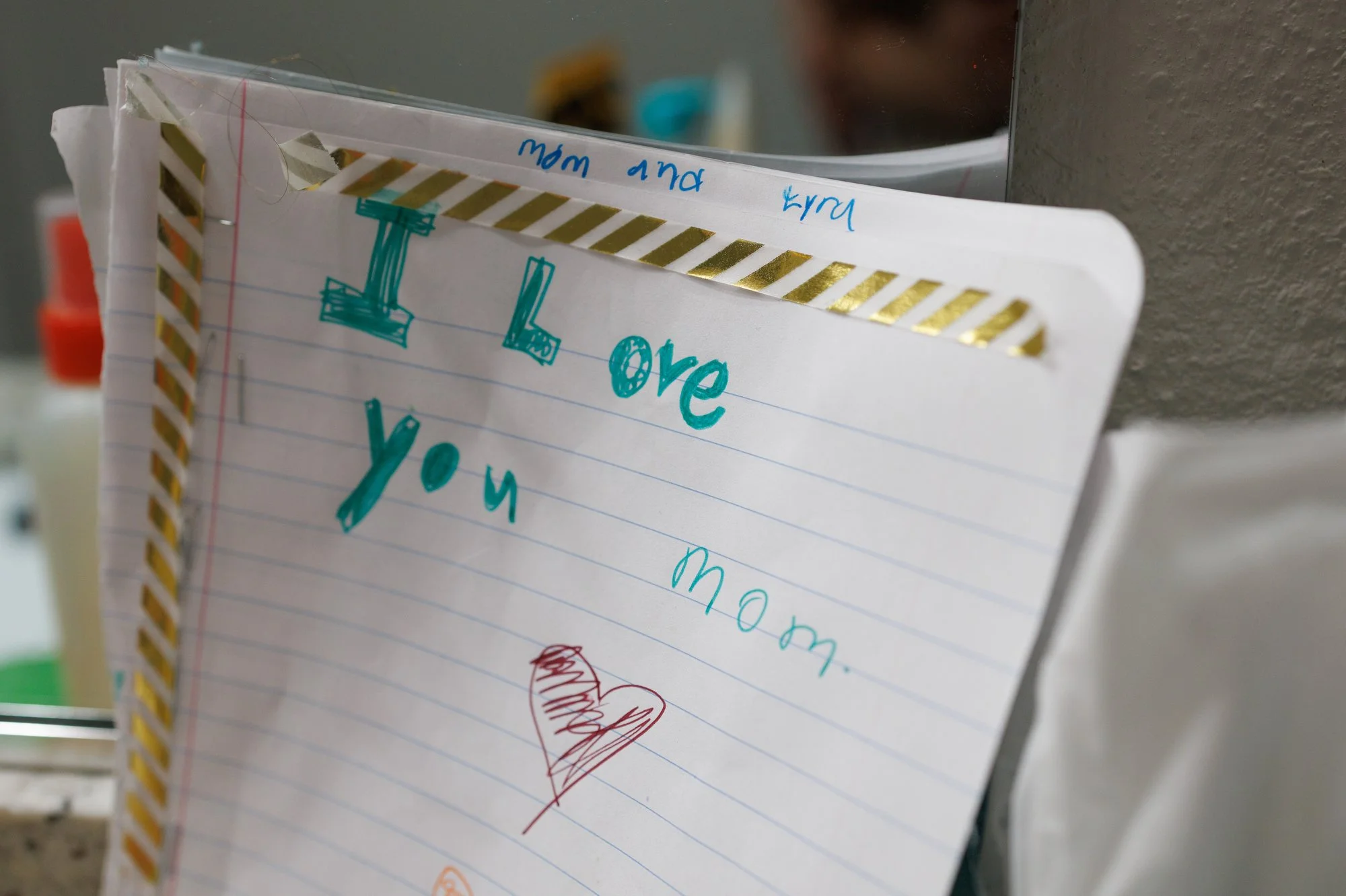 A piece of notebook paper with striped gold tape on the edges that has a child's block lettering that says "I love you mom" with a heart.