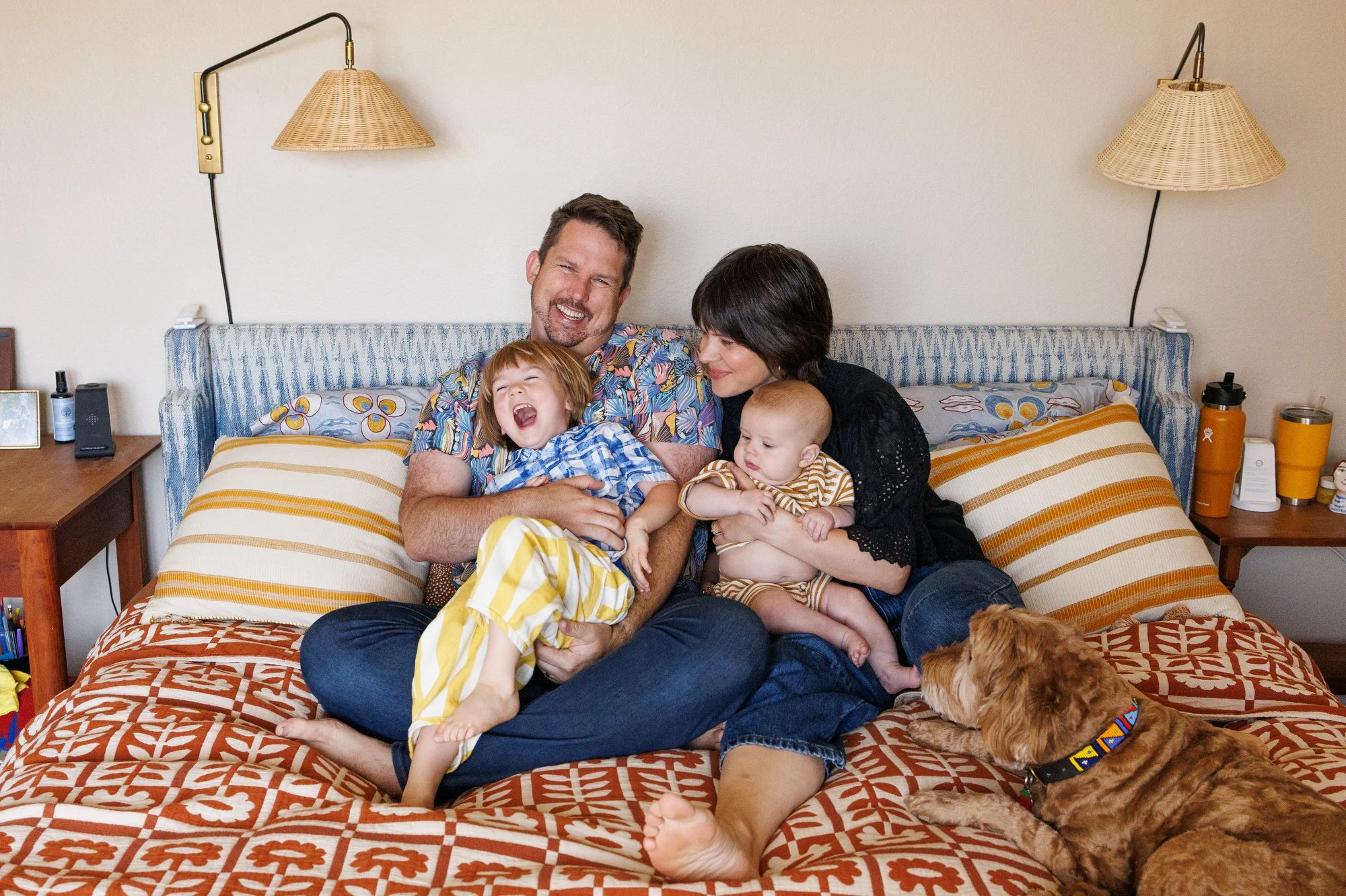 A family of four cuddles on the bed. Dad tickles son as both laugh. Mom holds an infant. A dog looks on. Photo by Shannon Fisher, an Orange County newborn photographer.