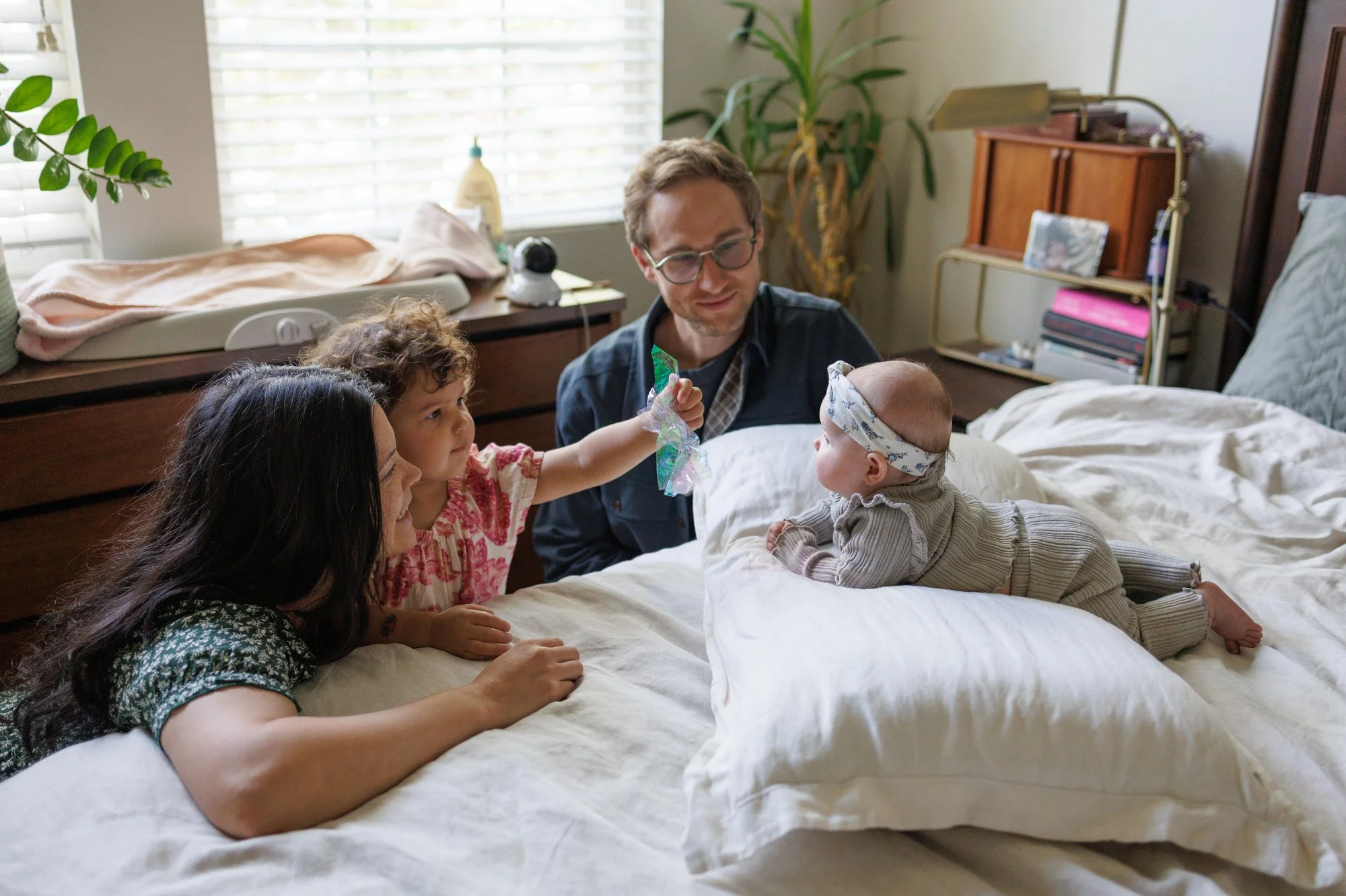 Newborn baby lays on her tummy on a pillow while her big sister holds shiny plastic up in front of her. Parents look on and smile.