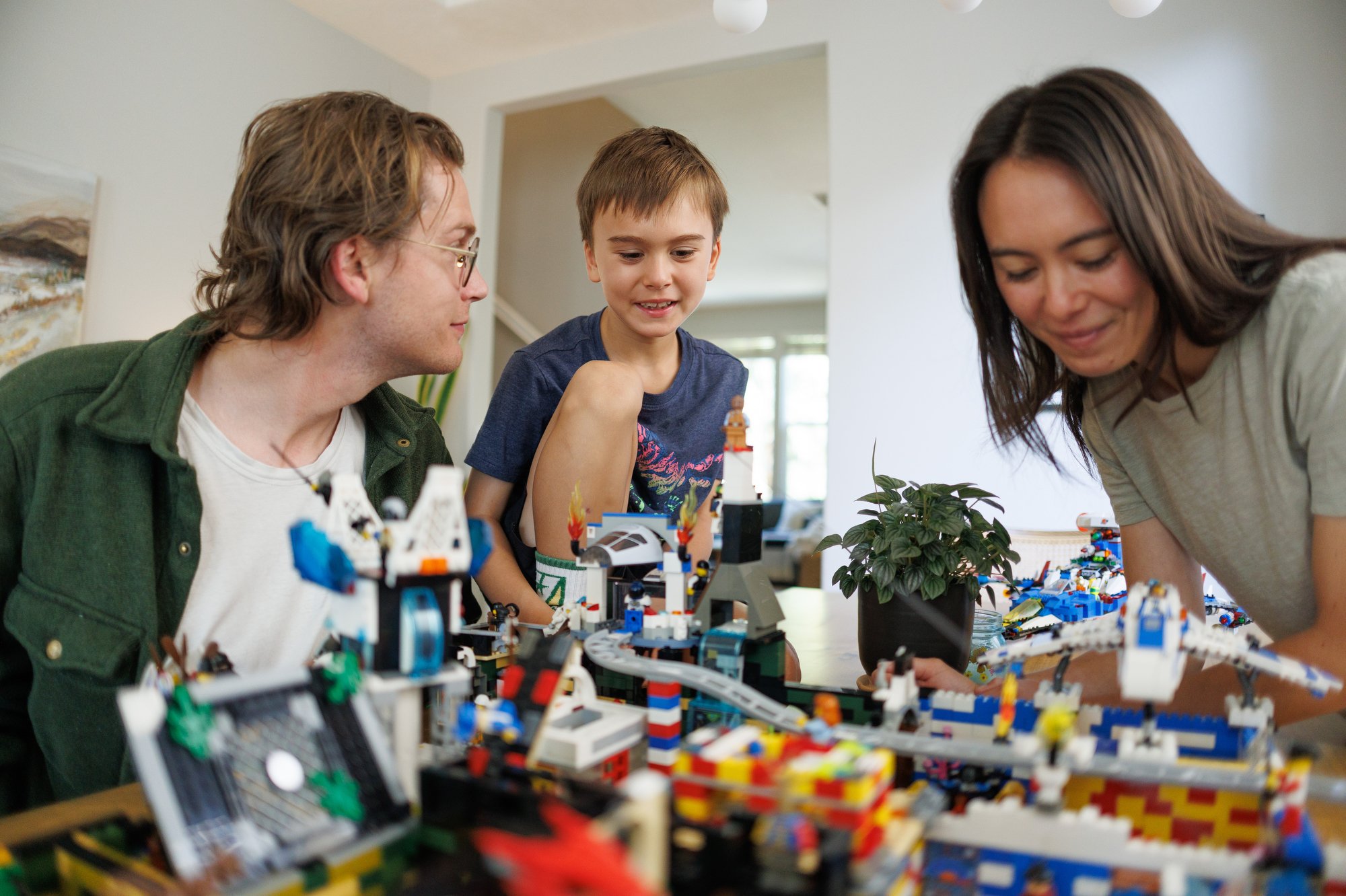 A 7-year-old boy sits on the dining table and looks excitedly at his Legos before him. His dad looks at him while his mom looks at the Lego creations.