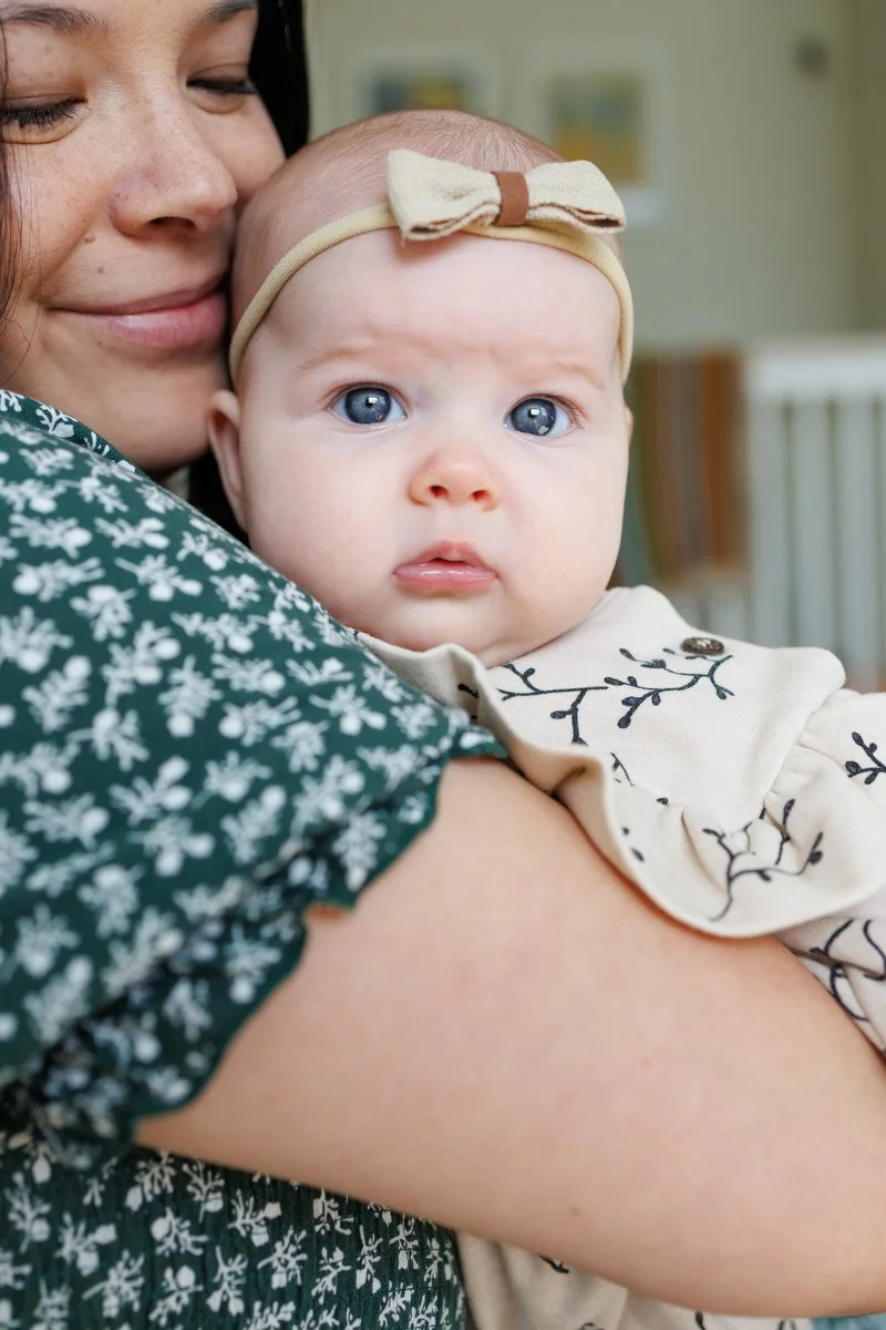 12-week-old baby with bow on her head looks into the camera with a furrowed brow and big blue eyes. Mom nuzzles her. Photo by Shannon Fisher.