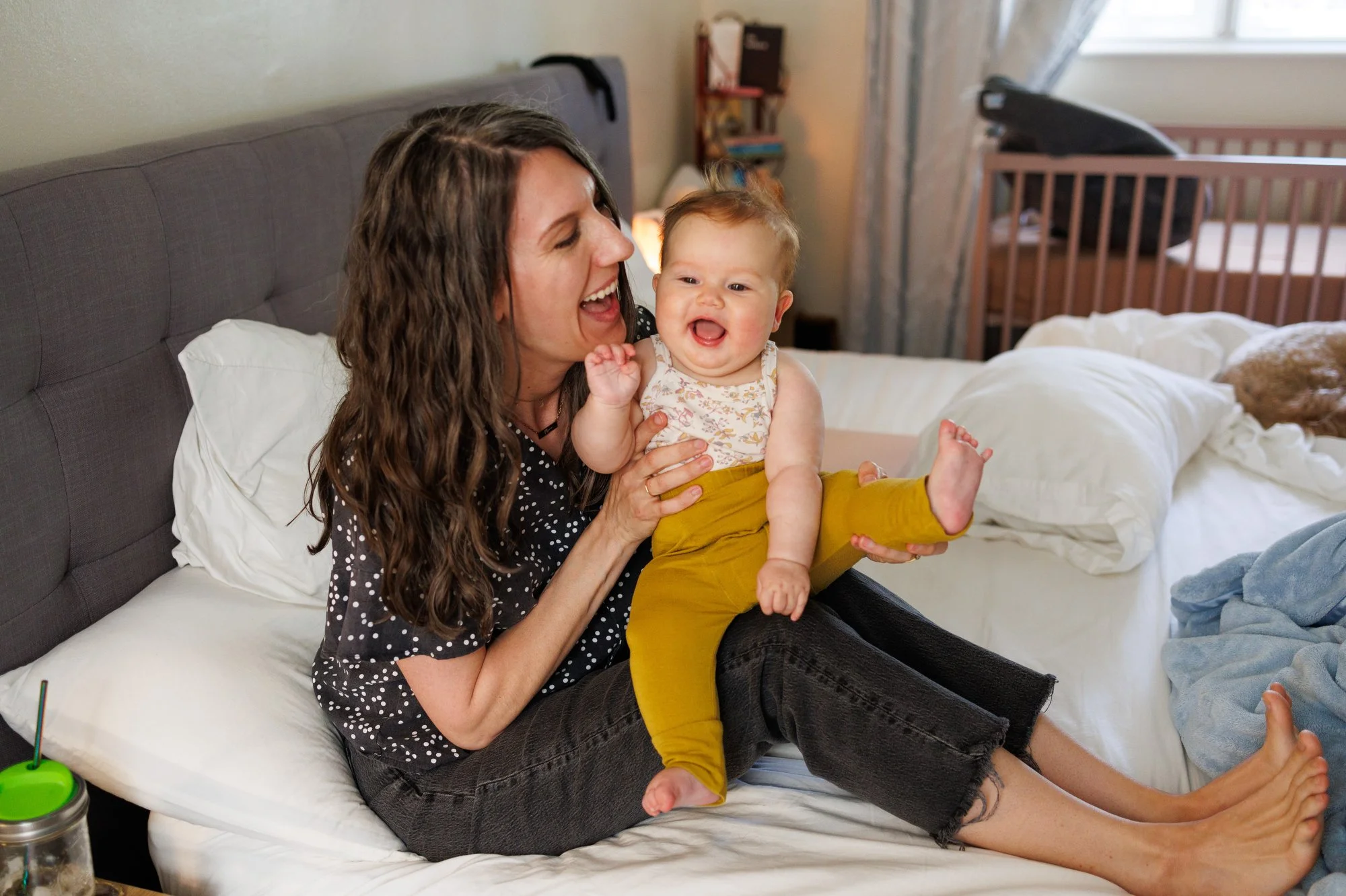 Photographer Shannon Fisher holds her baby on her lap on the bed, both have big smiles. Mom is holding baby's leg up in a high kick position.