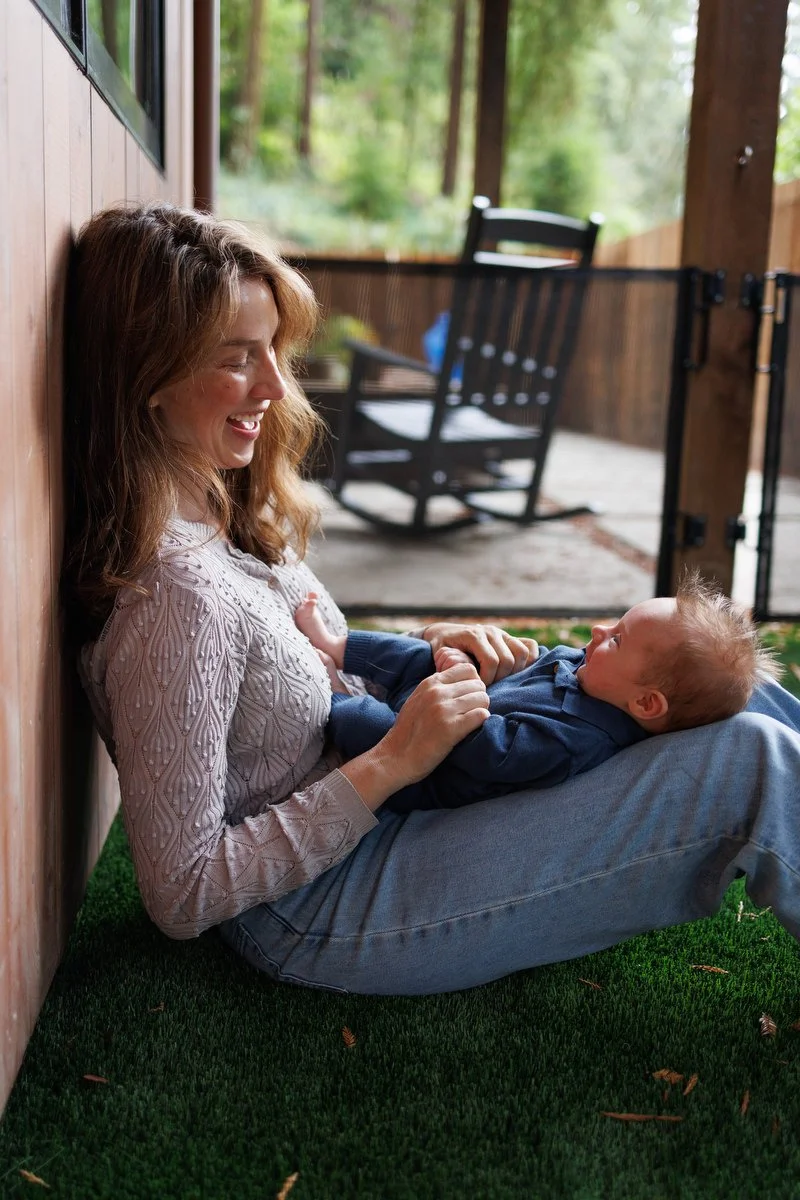 Mom sits leaning against the wall in her backyard holding her newborn baby in Orange County newborn photography session by Shannon Fisher Photo