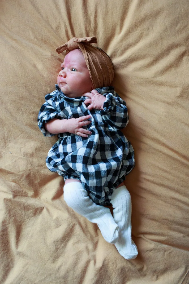 Tiny baby in a plaid onesie and tan headband laying on a bed with eyes open