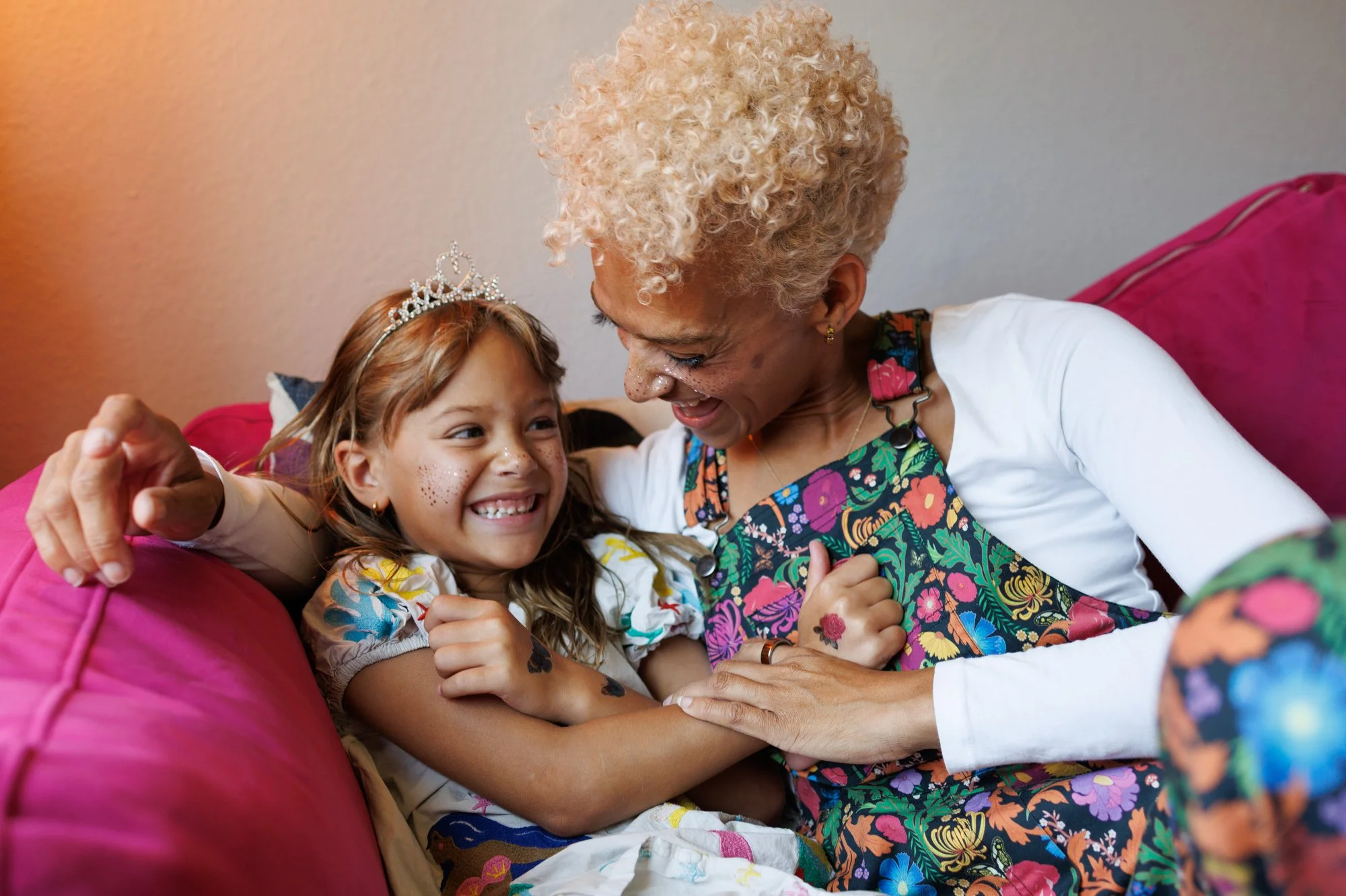 Average cost of family photos: A mom with floral overalls has her arm around her daughter, who is wearing a tiara and laughing.