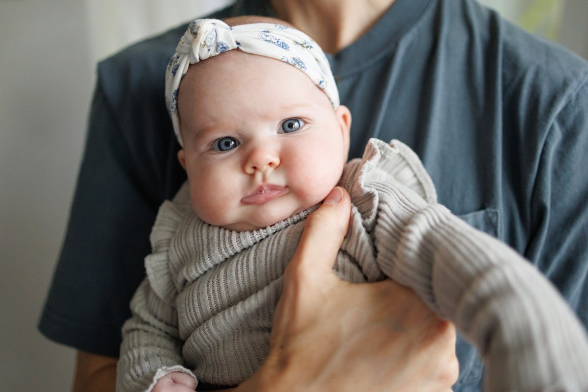 Baby with piercing blue eyes and white headband stares at the camera with a neutral expression. Her dad is holding her, but you just see his torso and his hand across her.