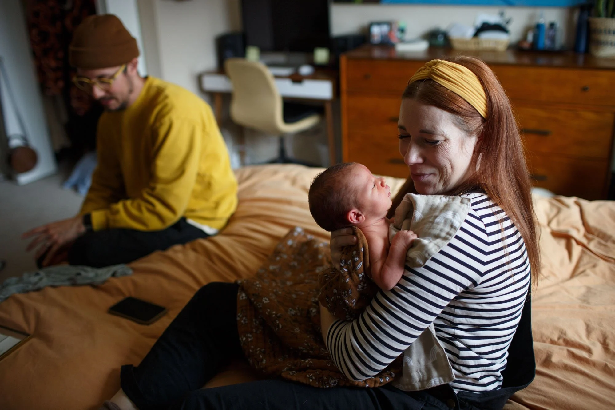 Mom smiles at her 1-week old baby wrapped in a brown blanket. Dad sits off to the side on the bed.