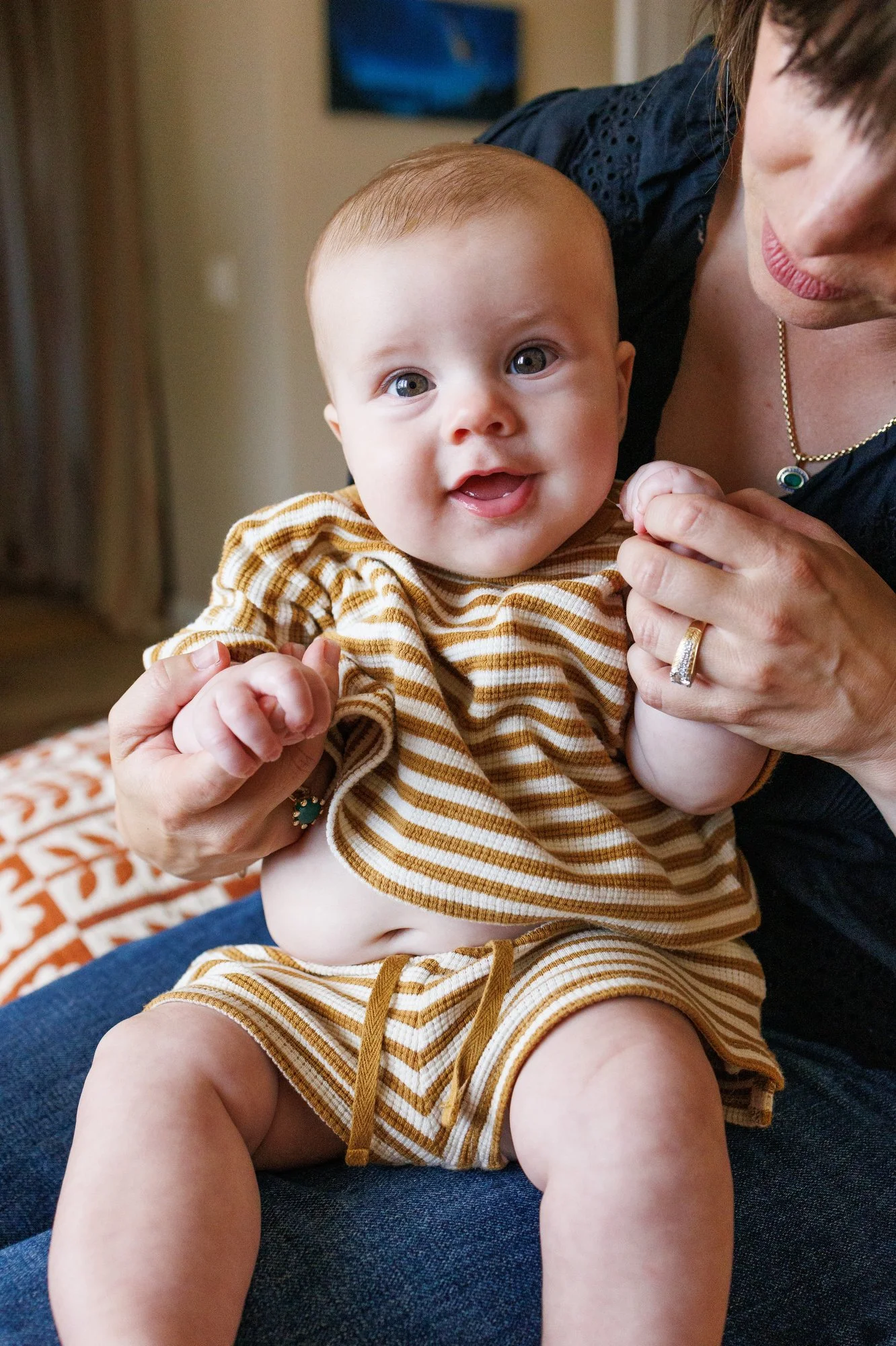 Baby with striped shorts and shirt smiles at the camera. His belly is poking out and he is holding onto Mom's finger as he sits in her lap.