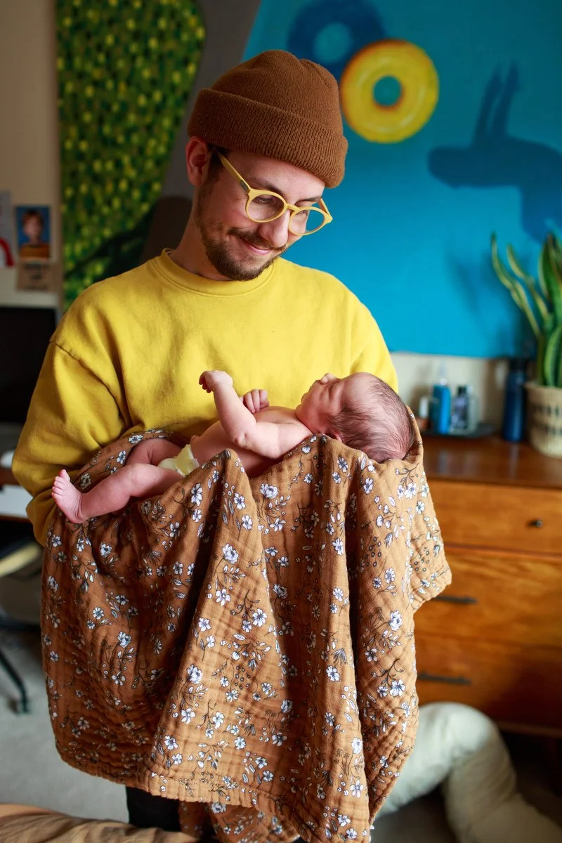 Dad in yellow glasses and matching yellow shirt smiles down at one-week-old daughter. Photo by Shannon Fisher, an Orange County newborn photographer.