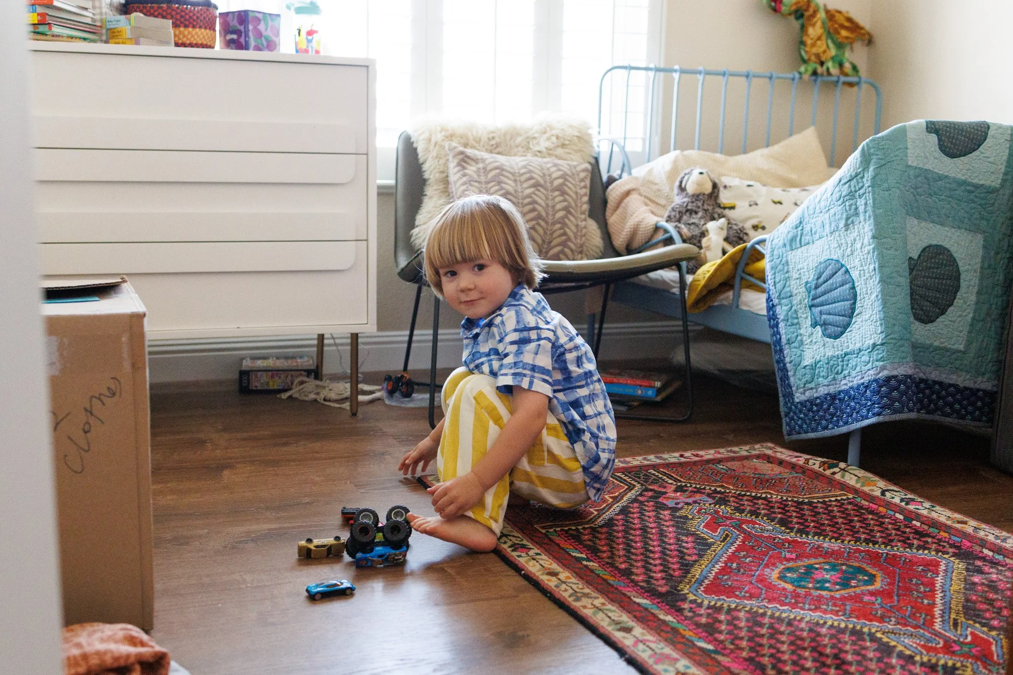 A little boy in yellow striped pants and a blue plaid shirt smiles at the camera as he plays with trucks in his room in an in-home family photoshoot by Shannon Fisher.