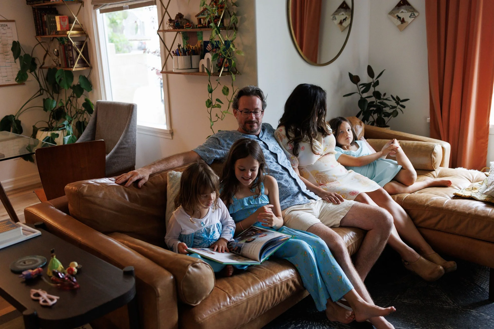 Mom and dad sit with three daughters on a leather sofa, reading. They're surrounded by plants. Photo by documentary family photographer Shannon Fisher.