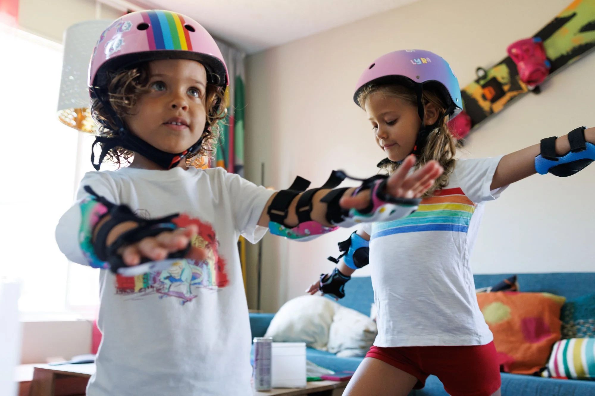 Two girls in rainbow shirts and rainbow helmets, wearing wrist guards and elbow pads, hold their arms out in their living room. In-home documentary-style photo session in Orange County, CA by Shannon Fisher.