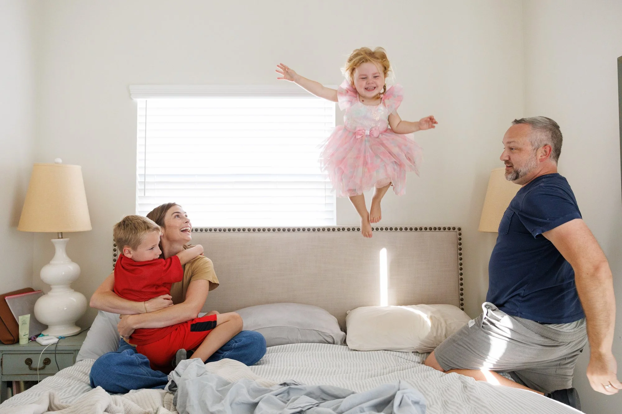 In a photo by OC family photographer Shannon Fisher, a little girl in a princess dress jumps high on the bed as her parents and brother watch