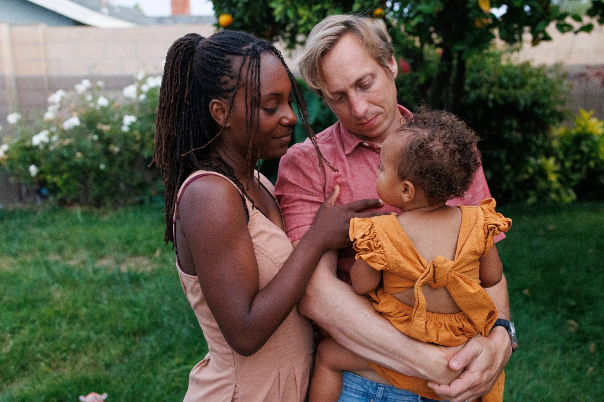 A family of three poses for their family photoshoot. Baby is in dad's arms in a golden orange ruffled outfit. Mom is in a warm light pink dress with her hand on the dad's chest. Dad is in a dusty coral shirt. Photo by Shannon Fisher.