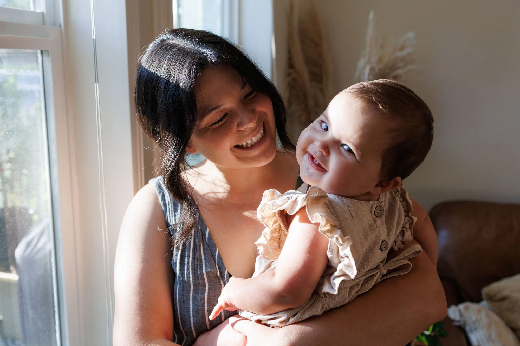 A baby turns over her shoulder and smiles at the camera while her mom smiles at her
