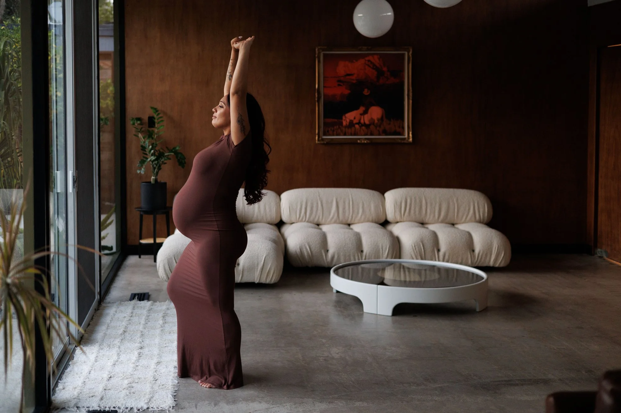 Pregnant woman in tight brown dress stretches arms above her head in a stylish midcentury living room. Photo by Shannon Fisher, maternity photographer in Orange County, CA.