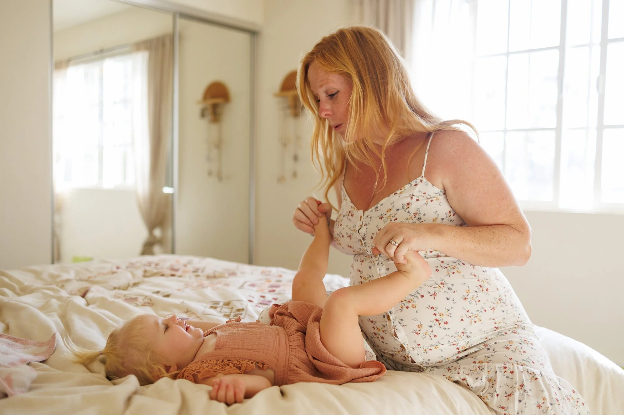 a pregnant mom in a white floral dress holds her little girl's feet as they sit on the bed