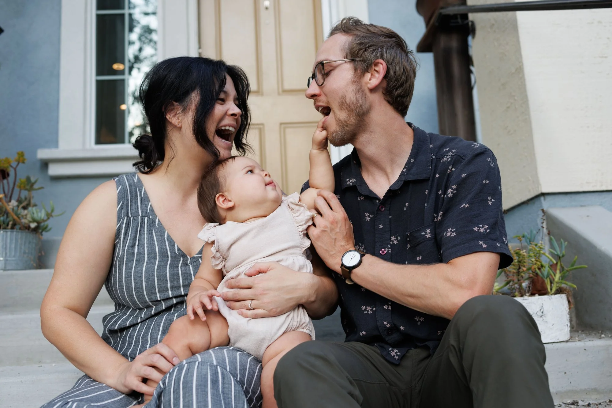 Family photoshoot outfit ideas: Mom and dad hold their baby on their porch. Baby is putting her hand in dad's mouth. Parents are in navy blue patterns, baby is in neutral ruffled onesie.
