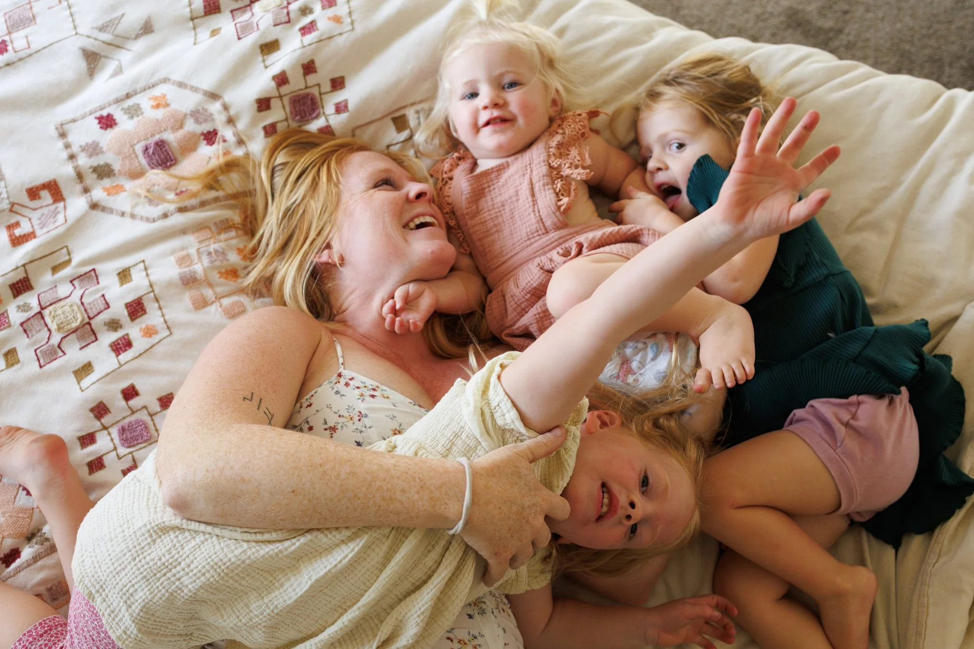 Mom lays on bed with her three daughters, laughing. All are in dresses: Mom in floral, girls in solid-color muslin ruffle dresses. Photo by OC family photographer Shannon Fisher.